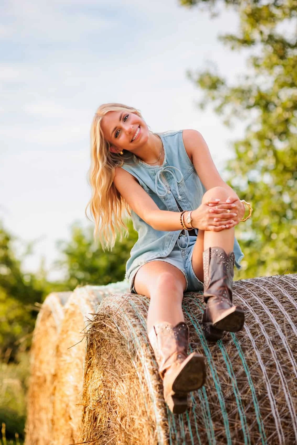  High school senior girl sitting on a hay bale outdoors, smiling, wearing a sleeveless denim top, shorts, and cowboy boots, with trees and a blue sky in the background.  Photo by Des Moines senior photographer Wendy Sorensen.