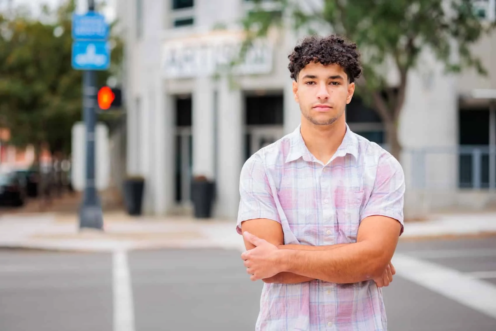  High school senior boy with curly dark hair and a light plaid shirt standing in the street in downtown Des Moines with arms crossed, looking at the camera, in front of a modern building with trees around.