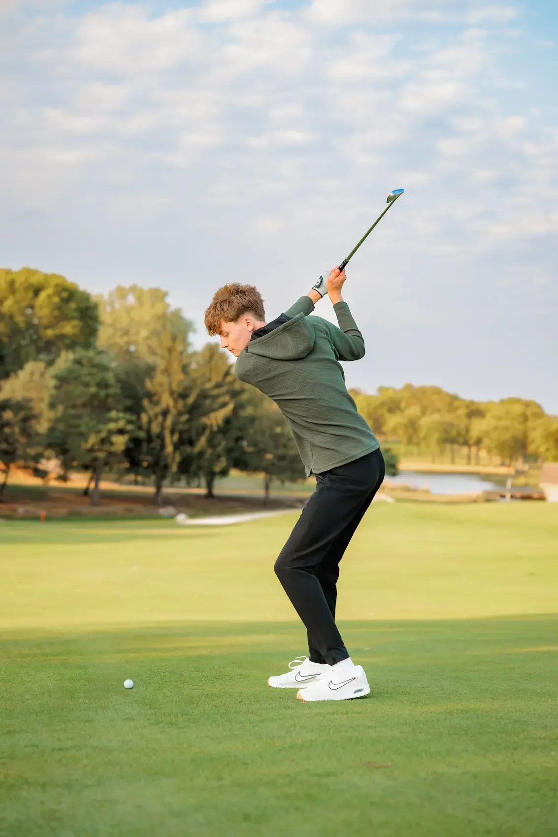 Senior boy playing golf on a golf course in Des Moines during his senior photoshoot, preparing to hit the golf ball with a club, surrounded by trees and an open sky.