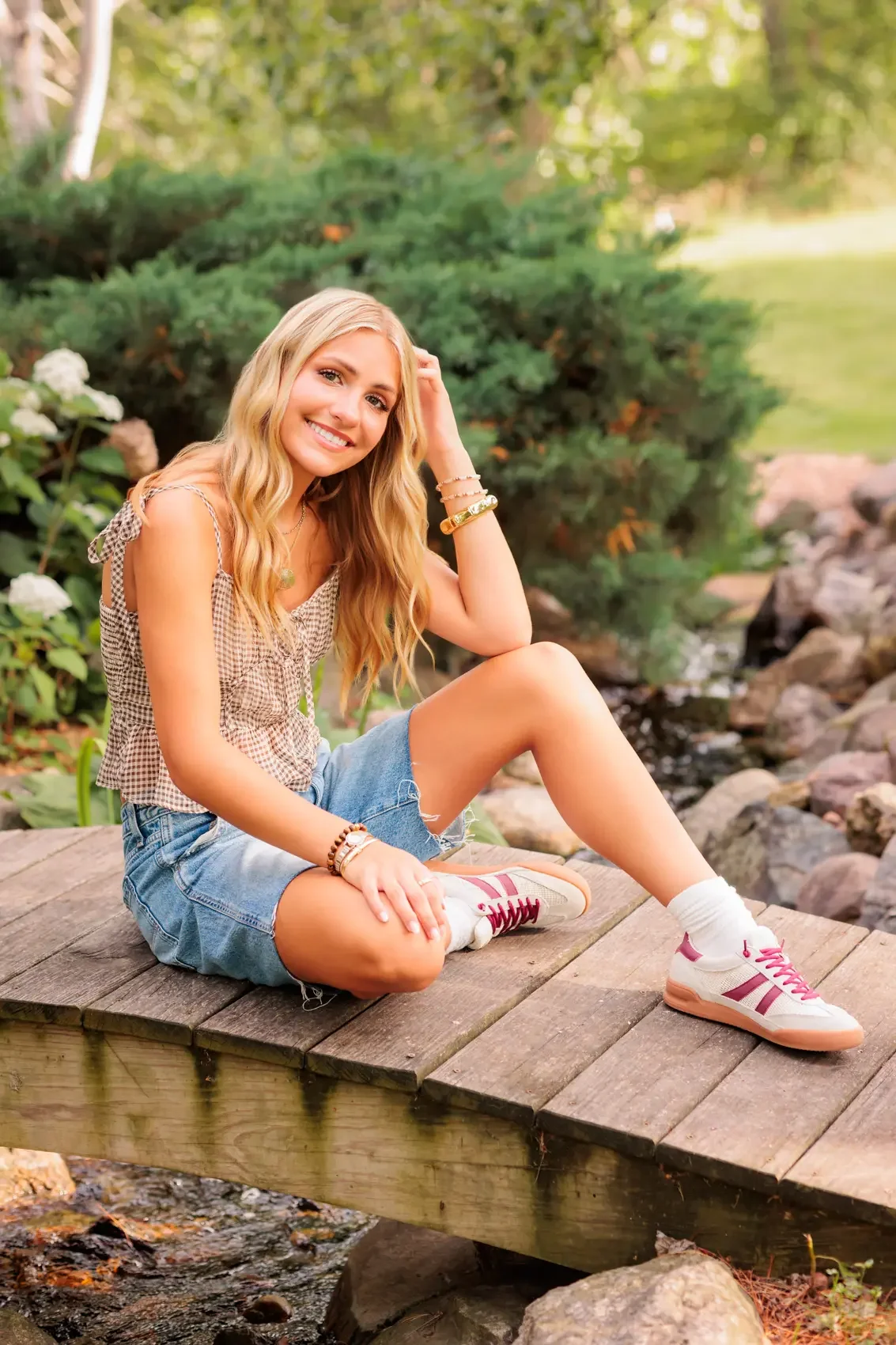 Senior girl with long, blonde hair sitting on a wooden bridge over a small stream in a lush garden, smiling at the camera.
