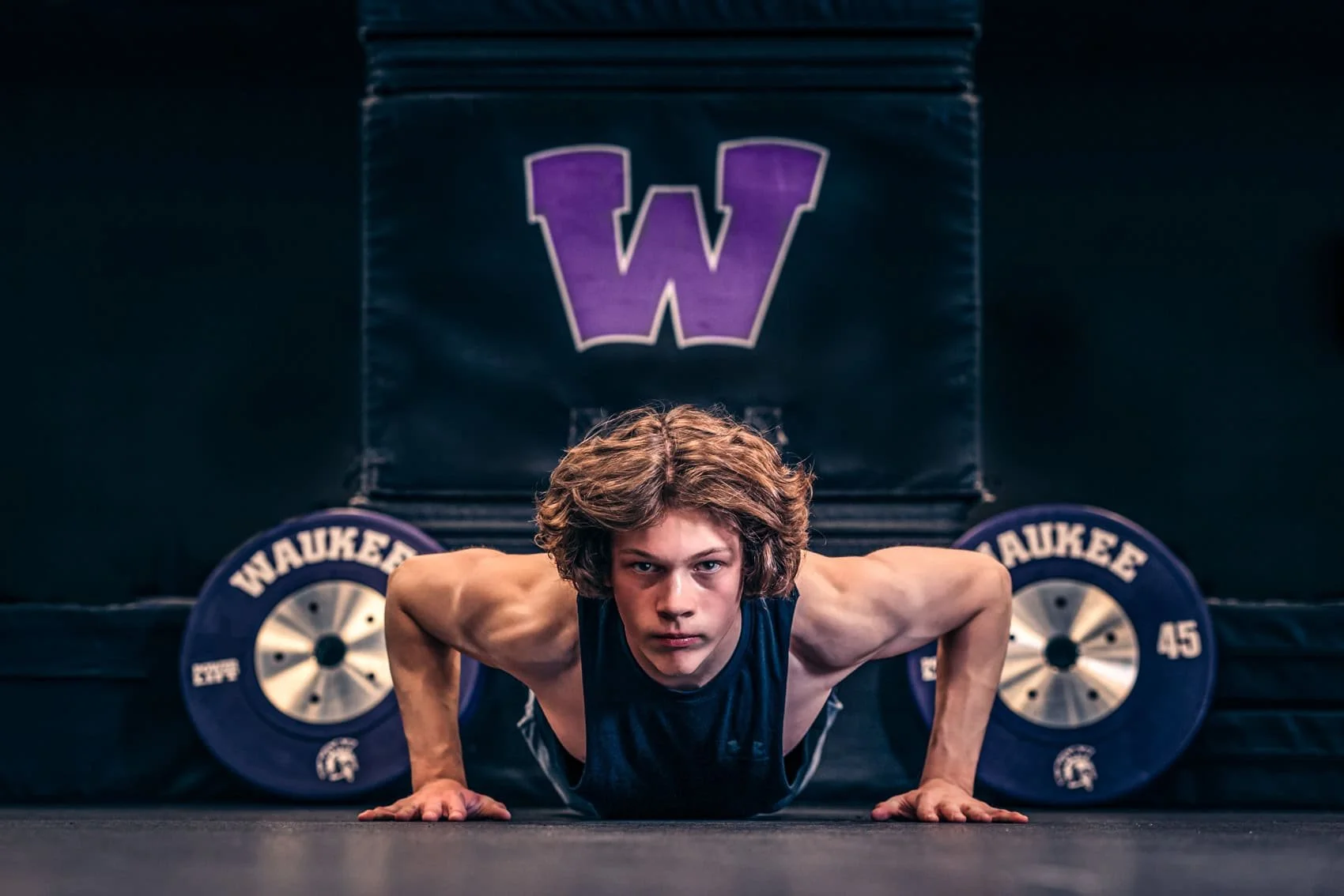  High school senior boy with brown, wavy hair doing a push-up in a gym with Waukee High School branding and weights in the background.