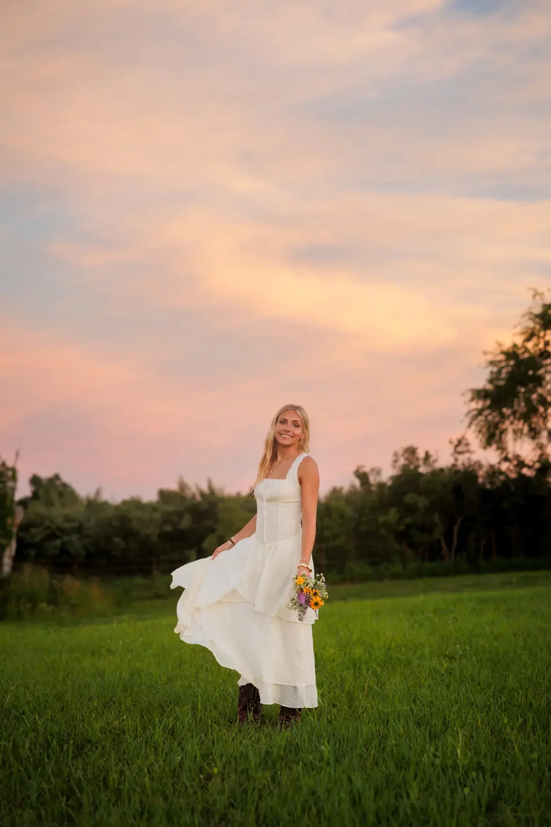Senior girl in a white dress holding a small bouquet of wildflowers in an open grassy field during sunset.