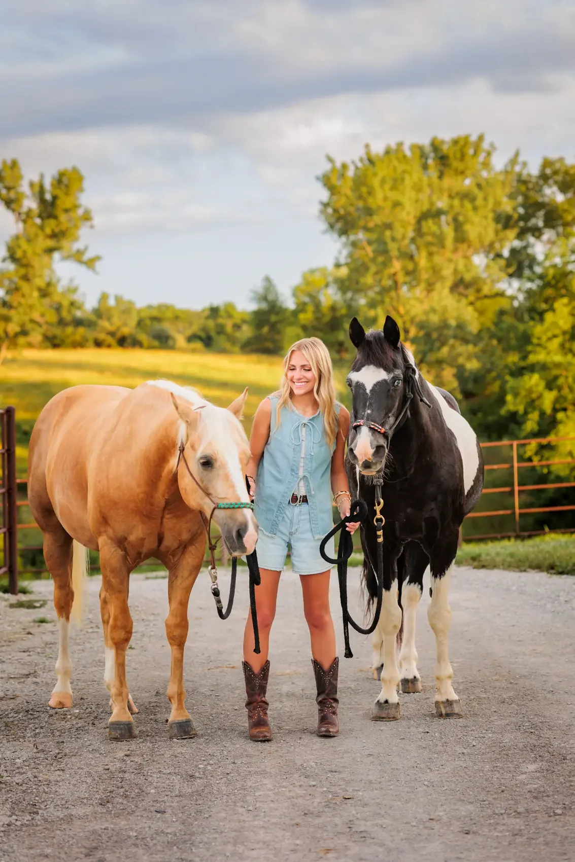 Senior girl standing between two horses outdoors on a farm, holding their reins, with greenery and trees in the background during her senior photo session.