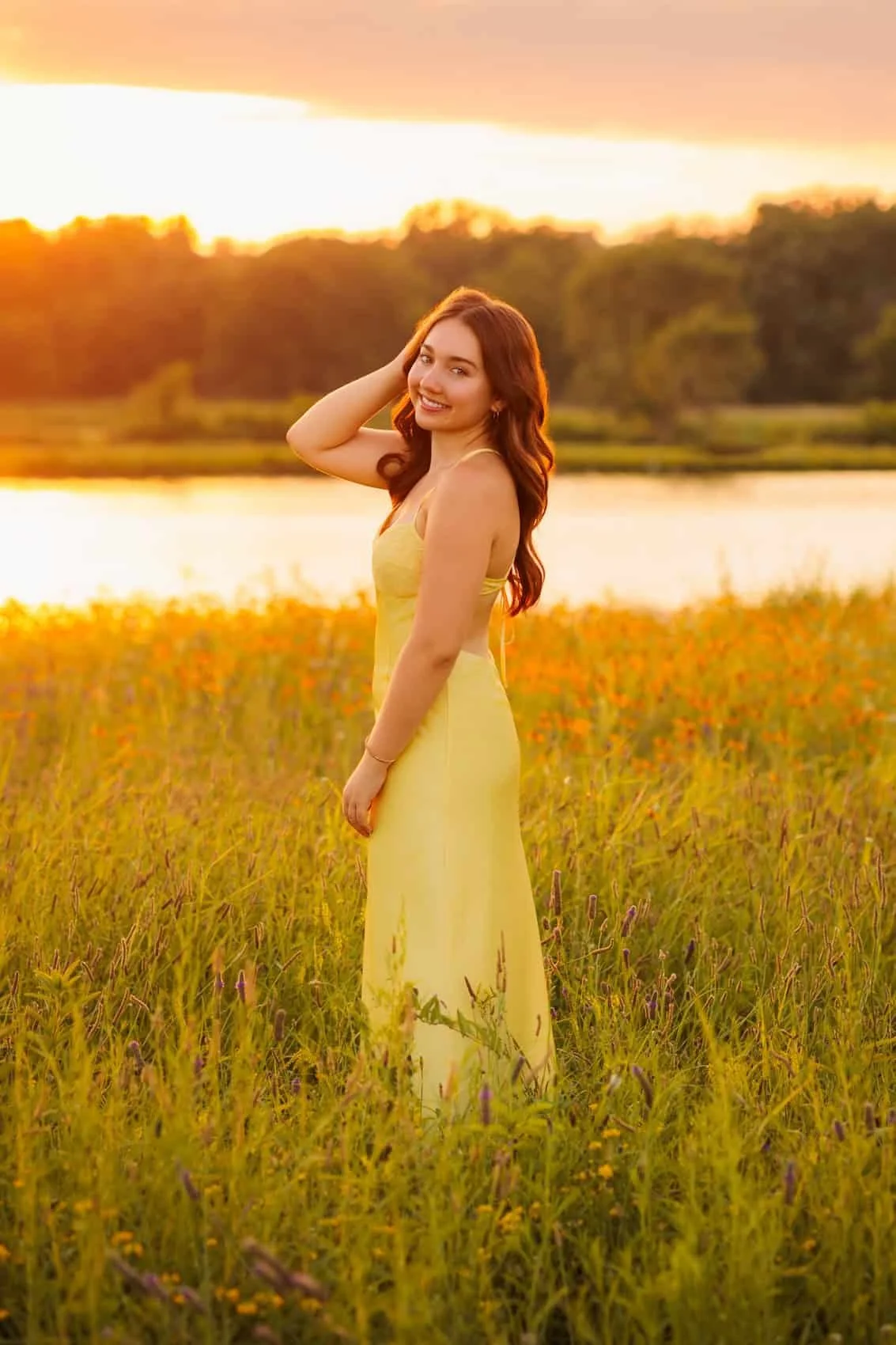  High school senior girl in a yellow dress standing in a field of wildflowers near a lake during sunset, smiling and looking at the camera. Photo by Wendy Sorensen, a Des Moines senior photographer.