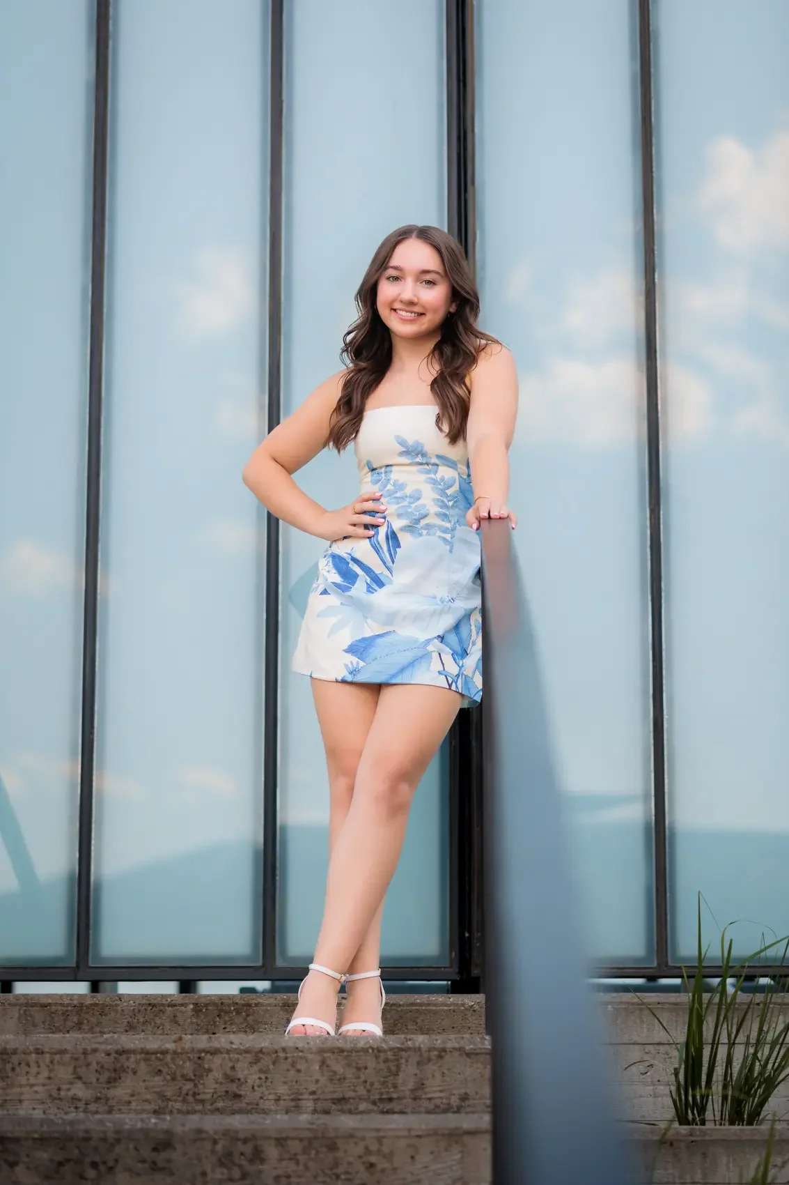 A young woman with long brown hair and a floral dress standing on steps in front of a glass building, smiling at the camera.