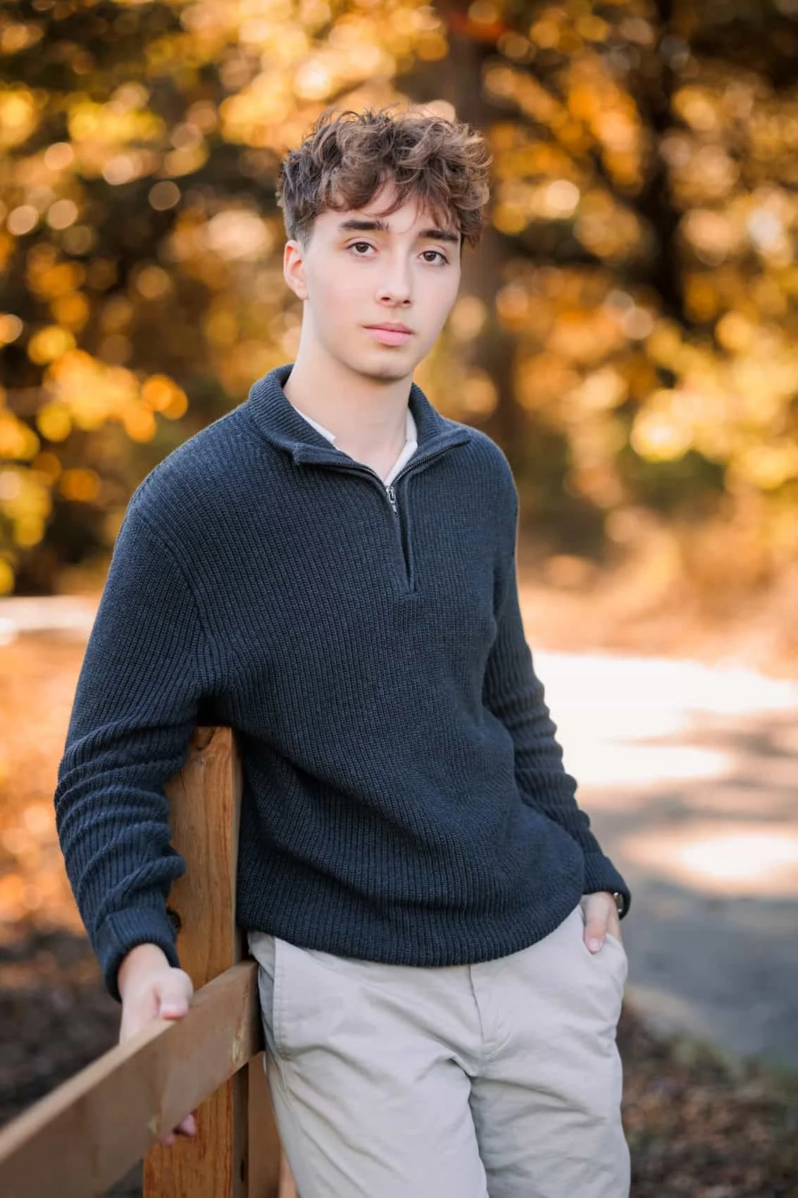  High school senior boy with curly brown hair, wearing a navy blue sweater and beige pants, stands outdoors in front of autumn foliage, leaning against a wooden fence.