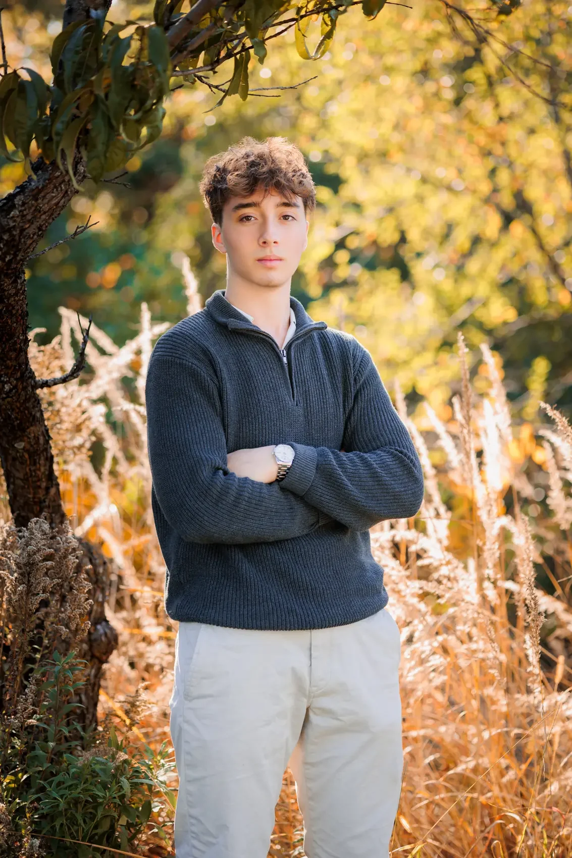 Senior boy with curly brown hair and light skin standing outdoors with crossed arms, wearing a dark sweater and beige pants, in a park with autumn foliage.  Image by Des Moines photographer Wendy Sorensen.
