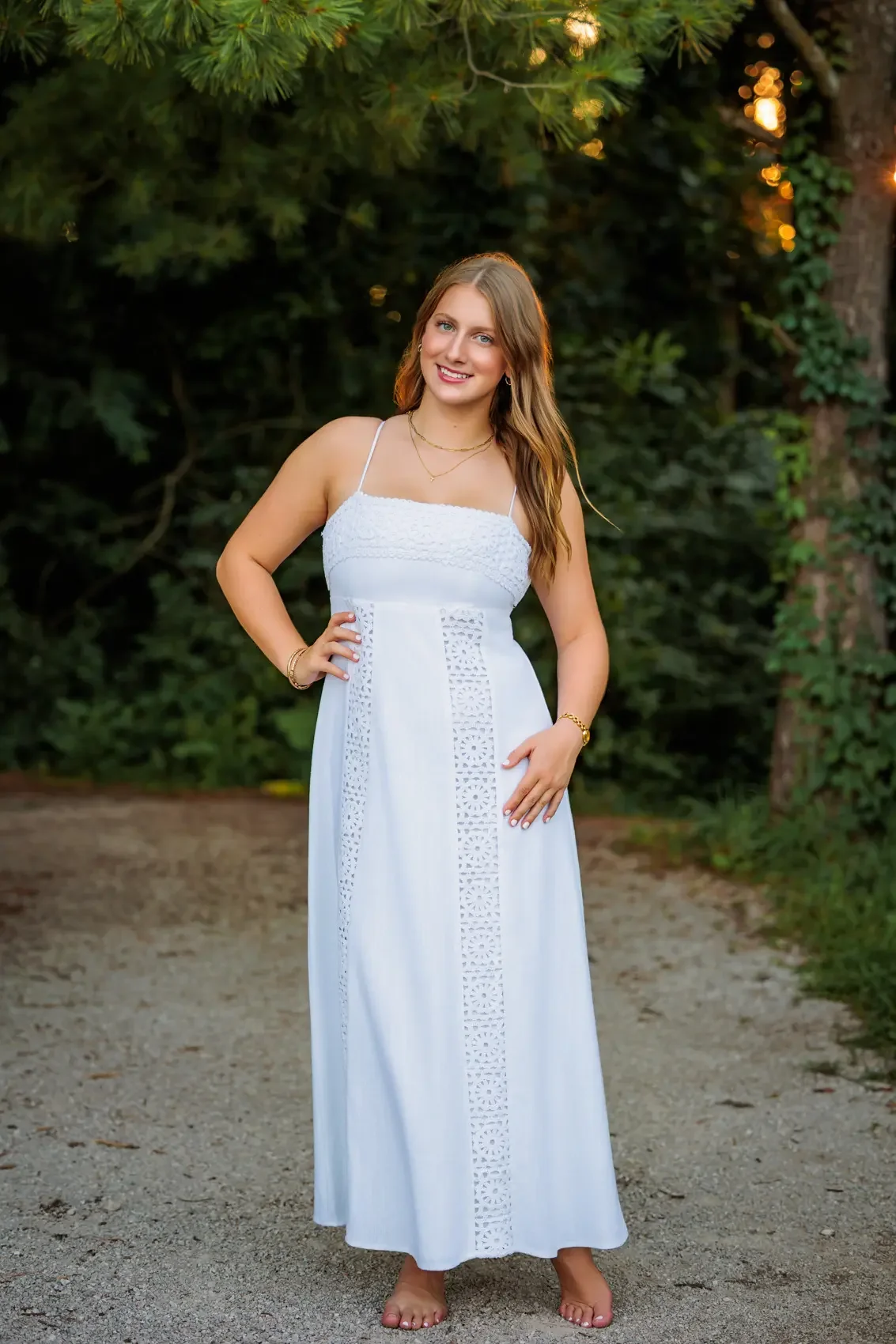 High school senior girl standing outdoors on a dirt path, wearing a white summer dress with lace details, smiling with her hand on her hip, surrounded by green foliage and trees.