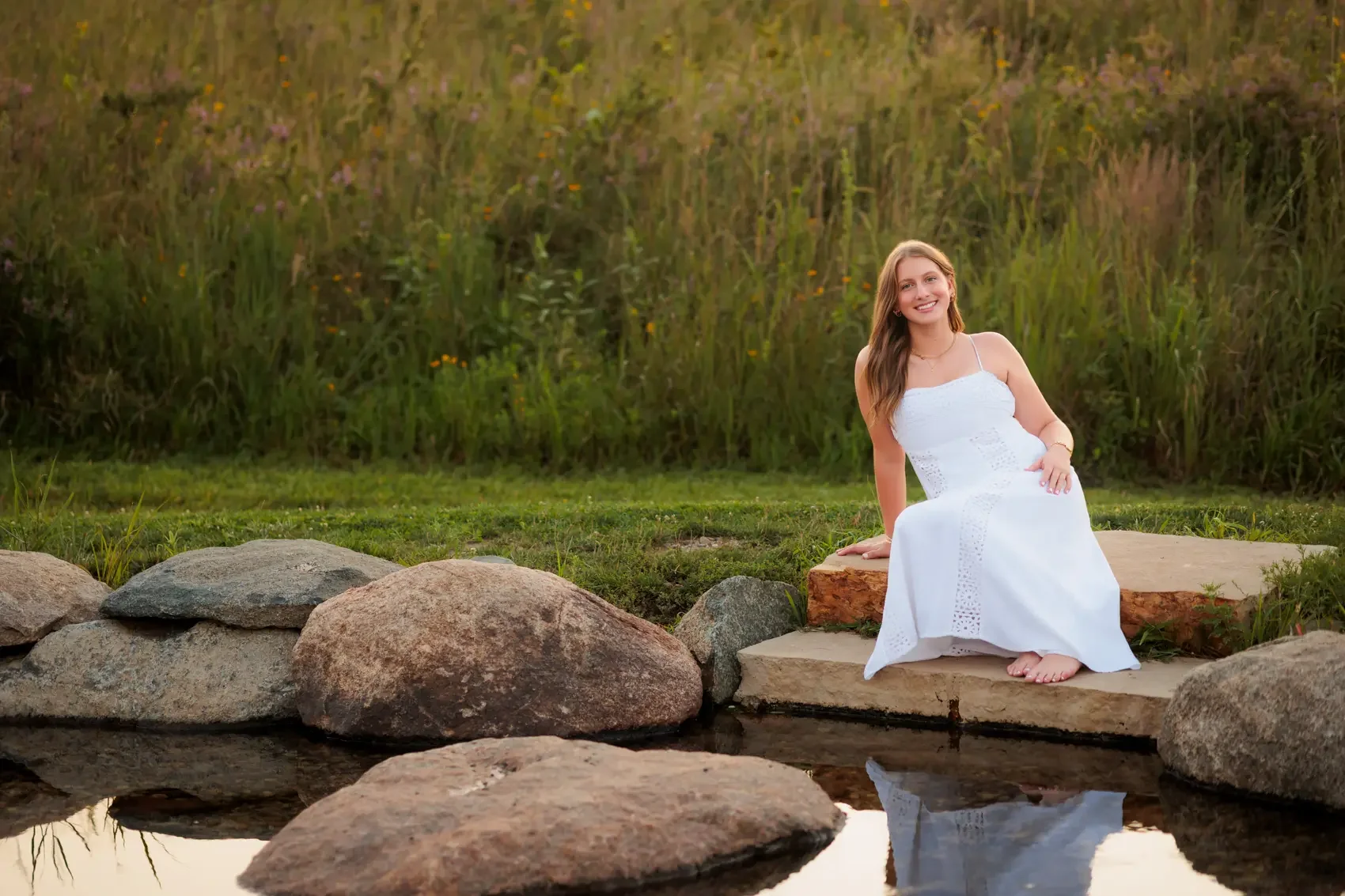A young woman in a white dress sitting on a rock near a small pond in a natural outdoor setting with greenery and tall grasses.