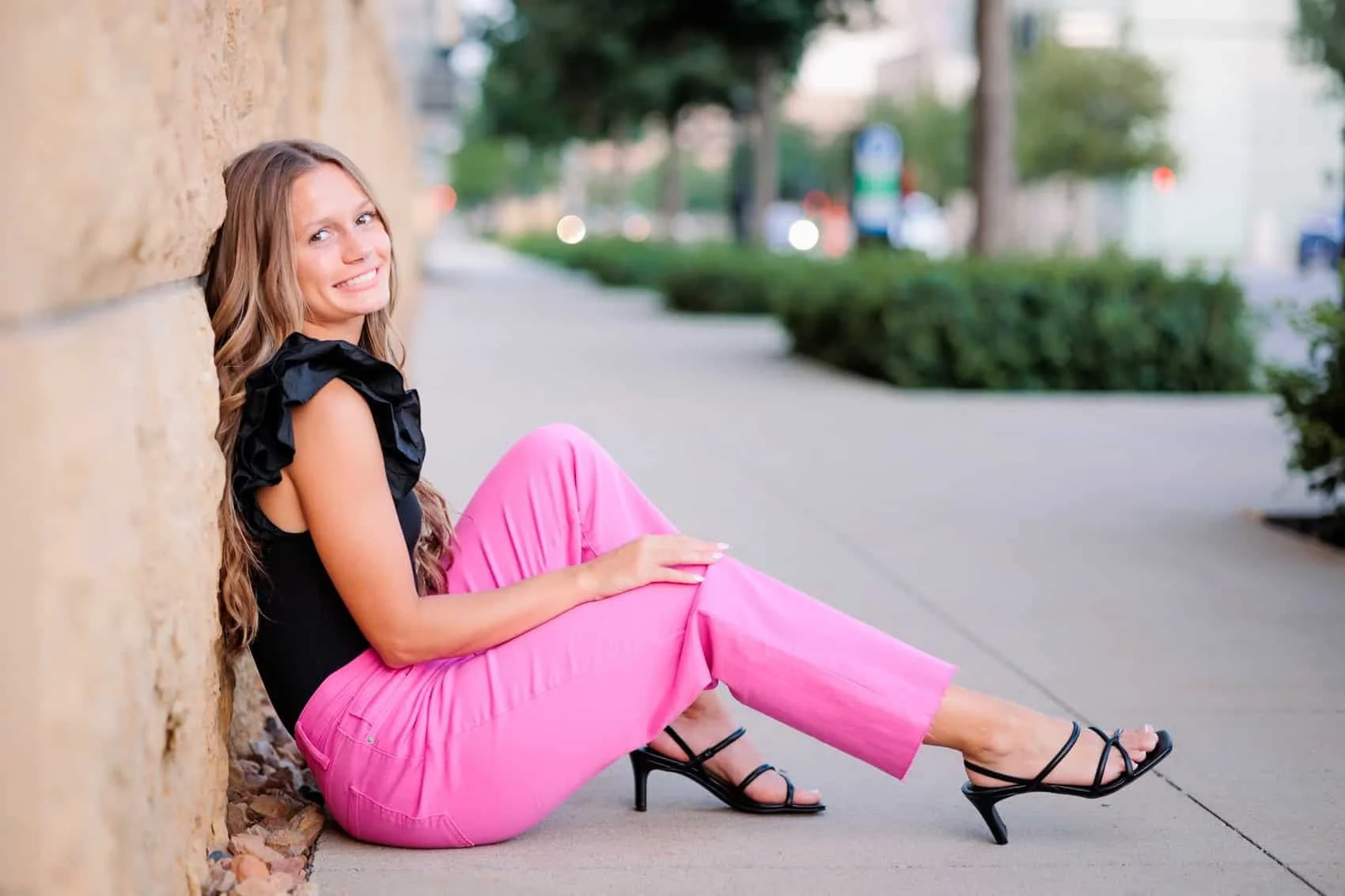 Senior girl with long, wavy hair sitting on the sidewalk in downtown Des Moines, leaning against a brick wall, wearing a black top with ruffled sleeves, pink pants, and black high-heeled sandals, smiling at the camera.