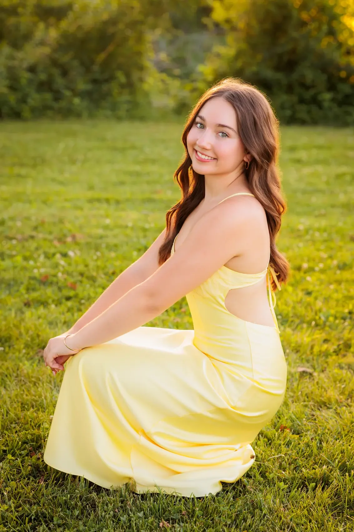 Senior girl with long brown hair, light skin, and blue eyes, sitting on grass in a park during golden hour sunset. She is wearing a yellow dress with thin straps, smiling at the camera with a gentle expression.