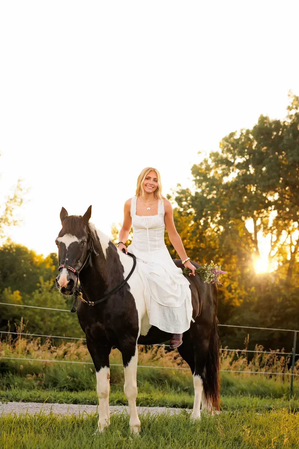 Senior girl in a white dress riding a black and white horse outdoors during sunset, holding a bouquet of wildflowers, with trees and a fence in the background.