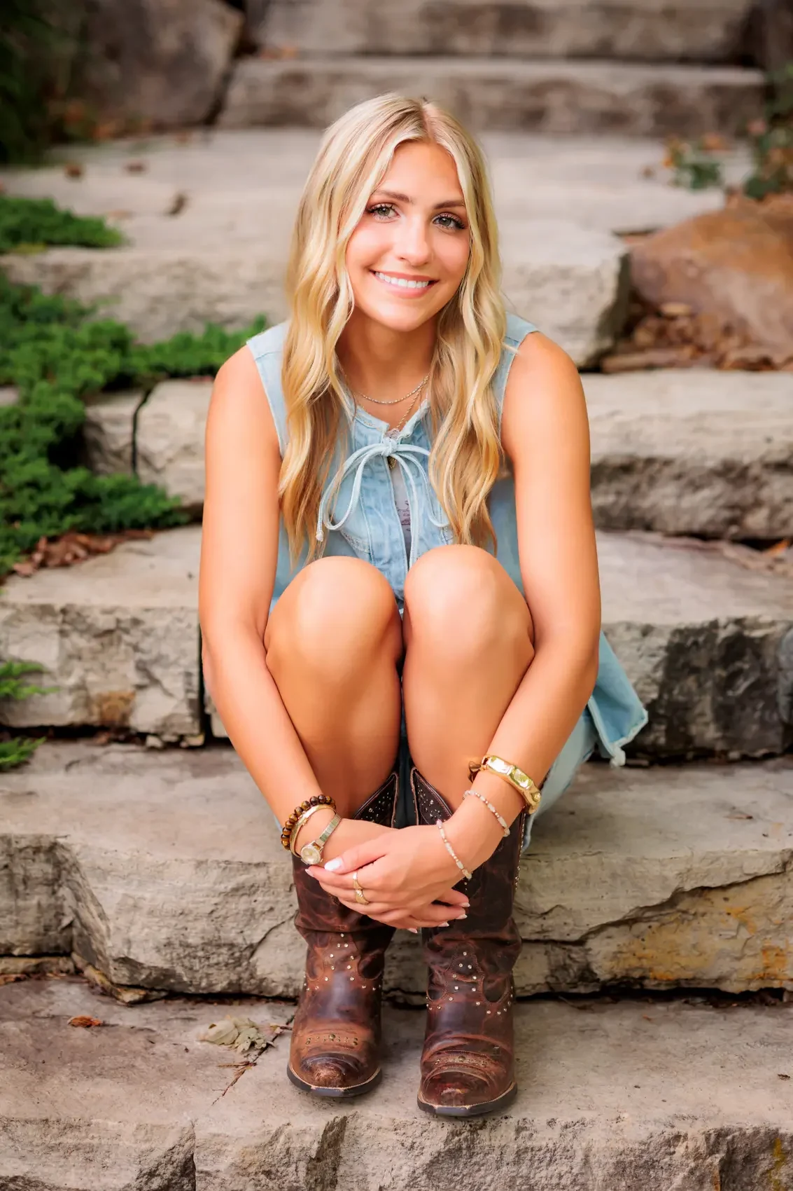 Senior girl with long blonde hair, wearing a sleeveless denim top, shorts, and brown cowboy boots, sitting on stone steps outdoors and smiling at the camera.