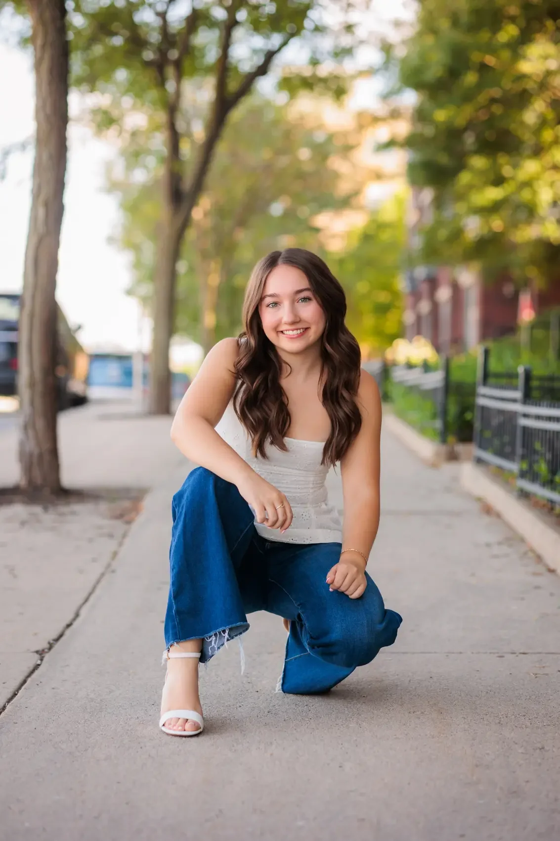 A smiling young woman with long brown hair kneeling on a sidewalk in a park or city street, wearing a white sleeveless top, blue jeans, and white high heels. Trees and buildings are in the background.
