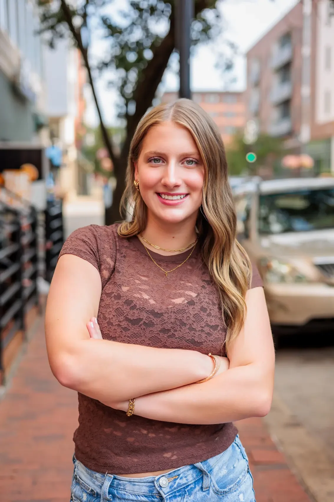 A young woman with wavy blonde hair and blue eyes standing outdoors on a city sidewalk, smiling with arms crossed, wearing a brown lace top and layered gold necklaces.