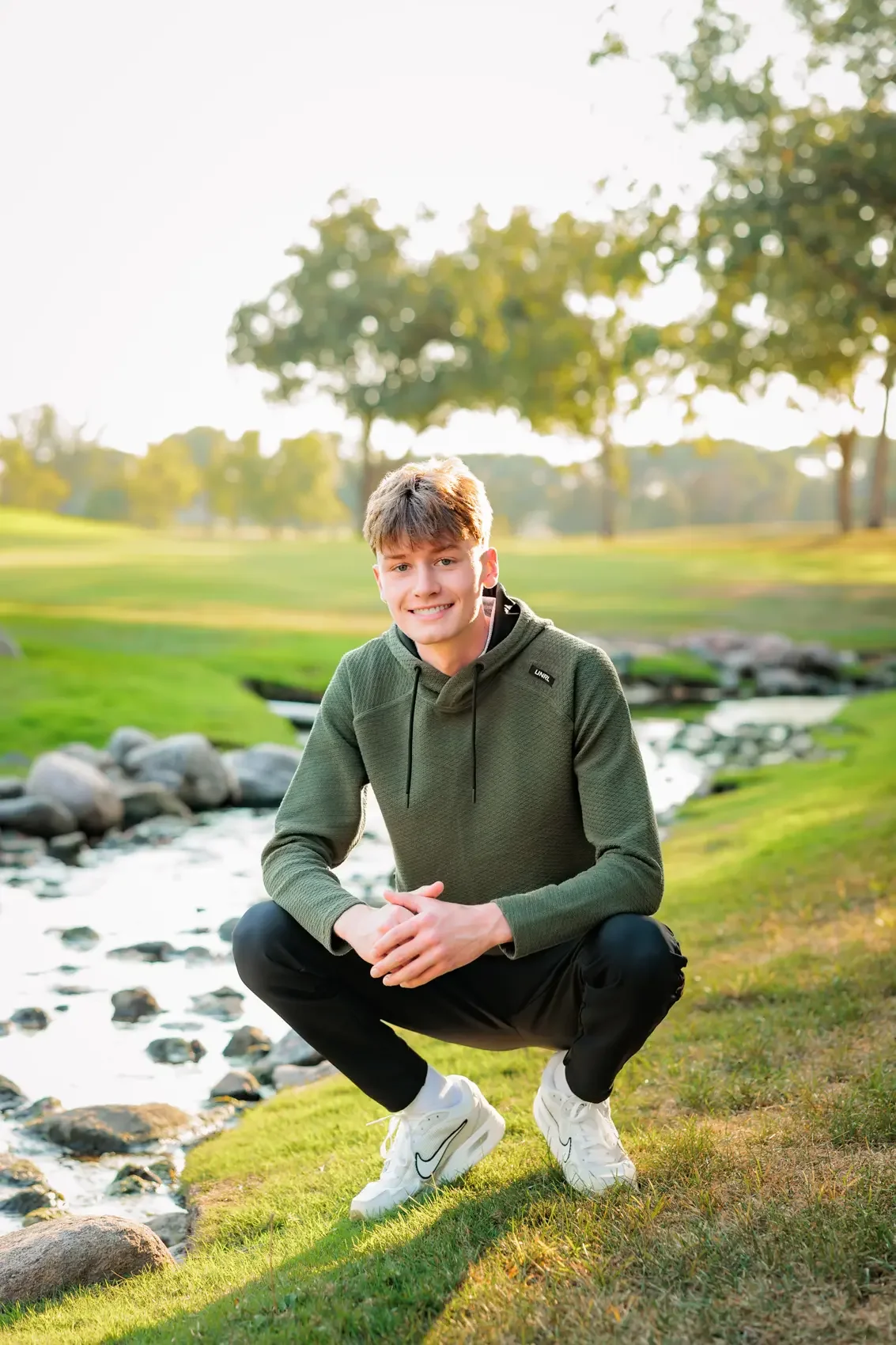 High school senior guy squatting on grass beside a small stream in a park with trees in the background, wearing a dark green hooded sweatshirt, black pants, and white sneakers, smiling at the camera.