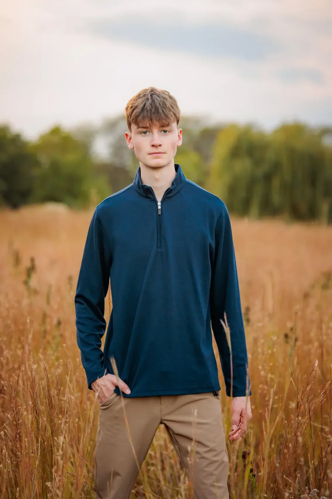 Senior boy standing in a field of tall grass, wearing a blue zip-up jacket and tan pants, with a background of trees and a cloudy sky. Photo by Des Moines senior photographer Wendy Sorensen.