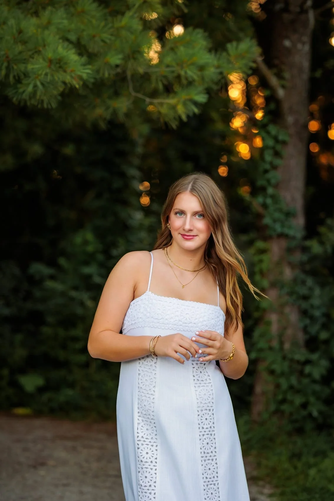  High school senior girl with long, wavy blonde hair is standing outdoors in a wooded area at sunset, wearing a white sleeveless dress with lace details, looking at the camera with a soft smile.