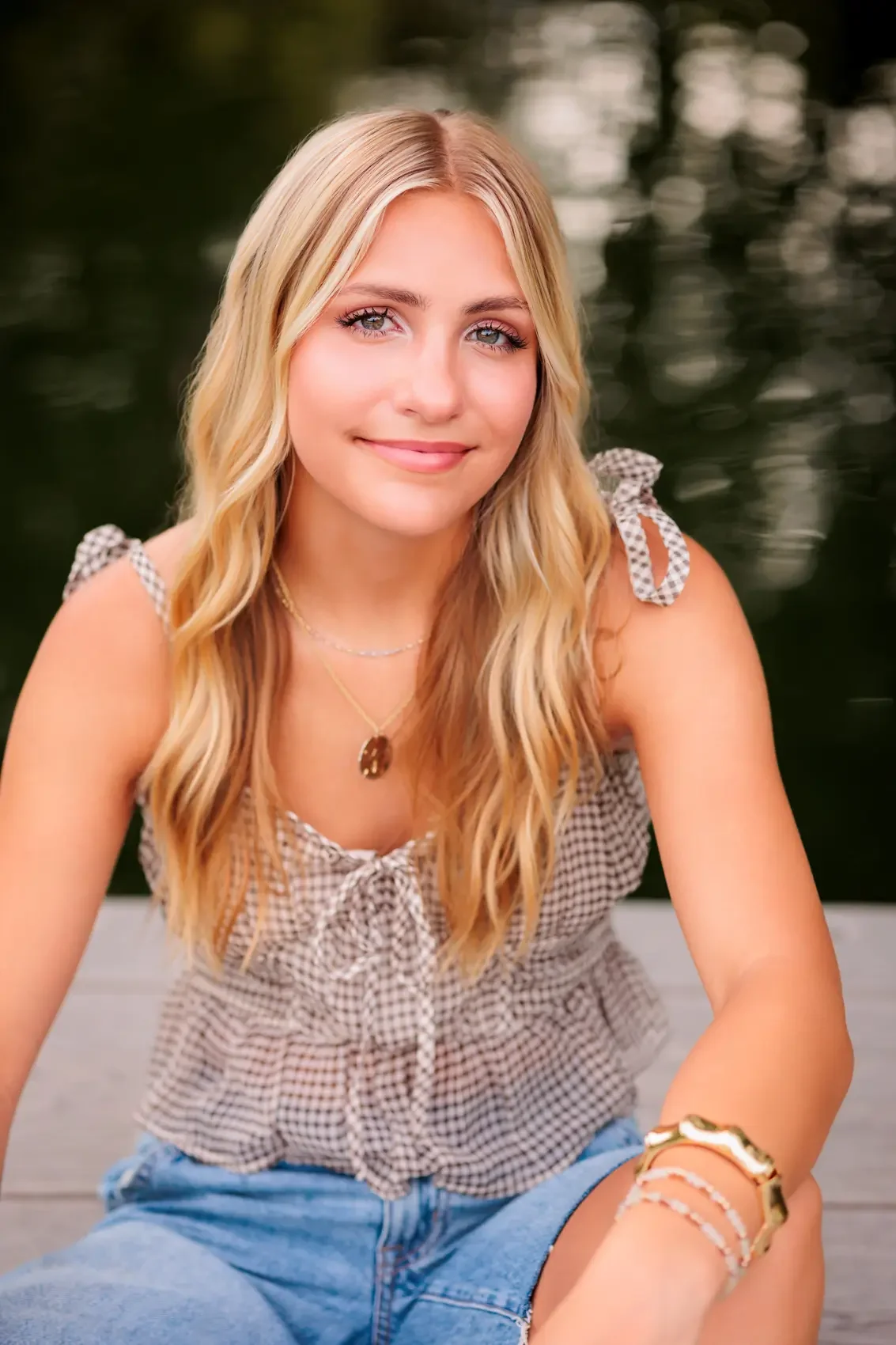 Senior girl with long blonde hair and blue eyes, posing outdoors near a body of water, wearing a checkered sleeveless top and denim shorts, smiling gently at the camera.