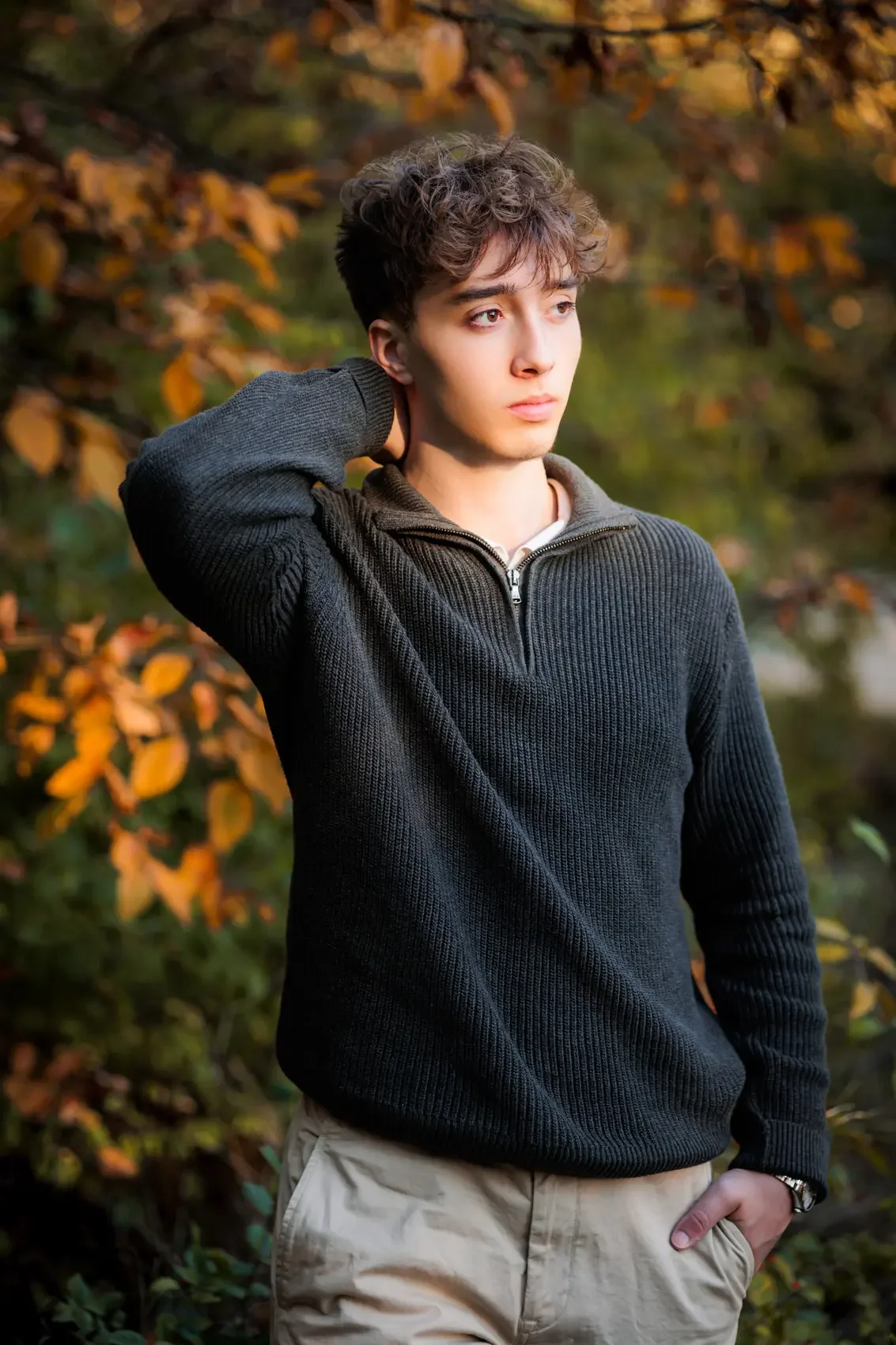Senior boy with curly brown hair standing outdoors among autumn foliage, wearing a black ribbed sweater with a half-zip collar and beige pants, looking thoughtfully into the distance. Photo by Wendy Sorensen, a Des Moines senior photographer.
