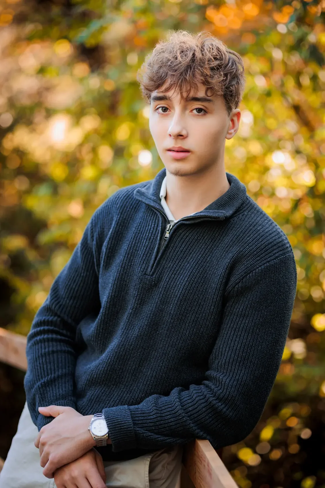 High school senior guy with curly brown hair and fair skin leans on a wooden railing along a Des Moines trail for senior pictures, wearing a navy blue zip-up sweater and a watch, with a background of autumn-colored foliage.