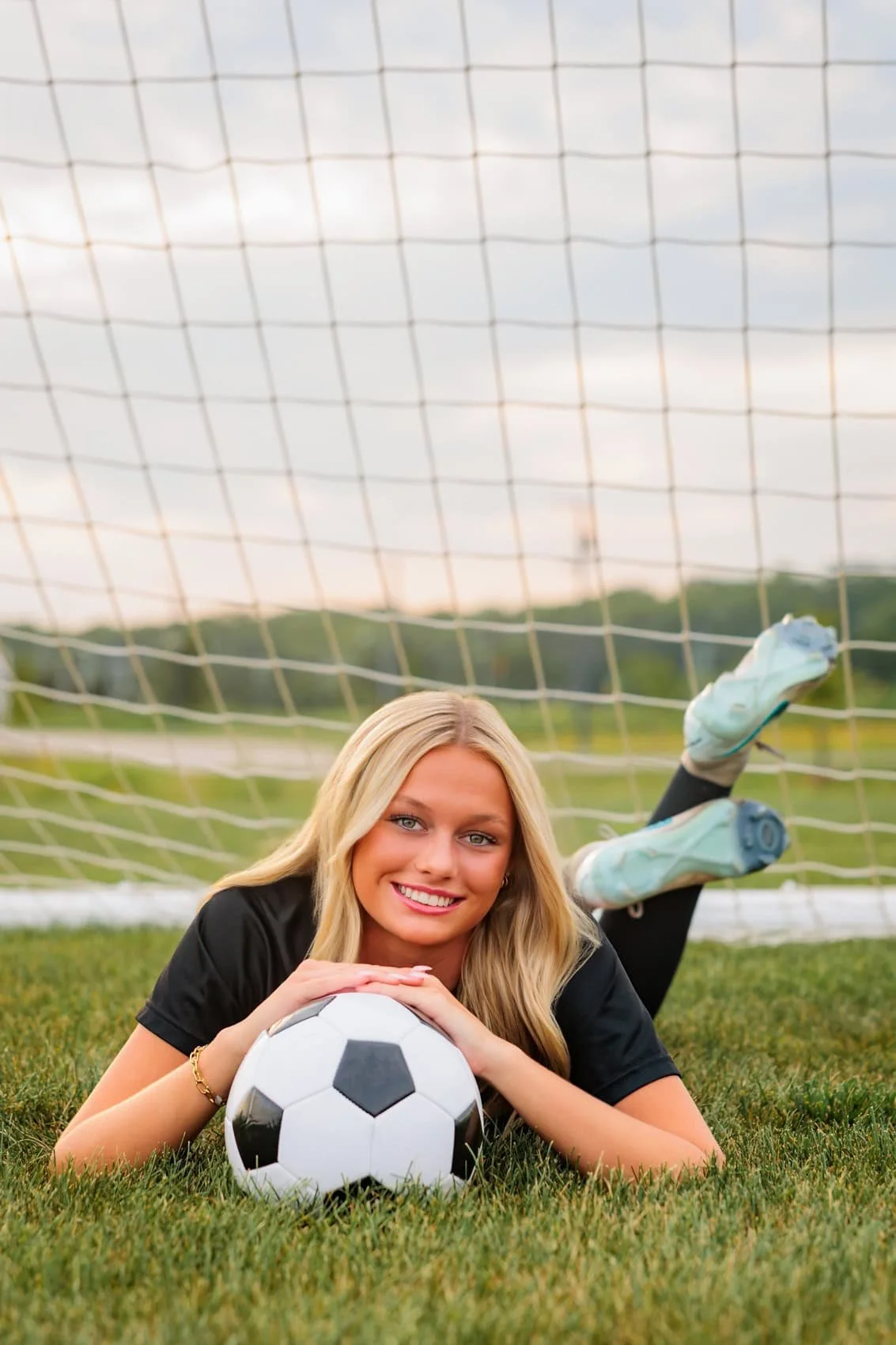 A smiling young woman in a black sports jersey lying on the grass with her arms resting on a soccer ball, in front of a soccer goal, during the daytime.