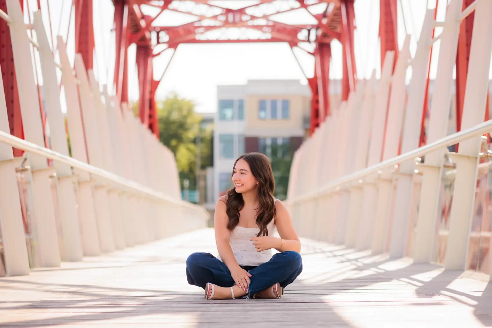 High school senior girl sitting cross-legged on a bridge, smiling and looking to her left, with trees and buildings in the background in downtown Des Moines.