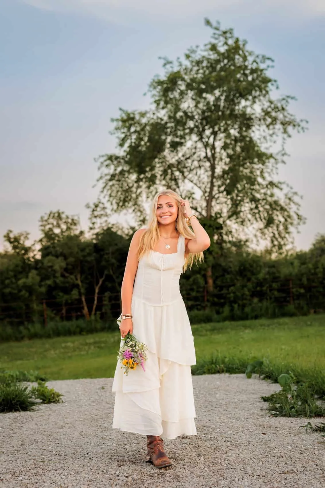 Senior girl in a white dress holding a small bouquet of flowers, standing outdoors on a gravel path with greenery and a tree behind her, smiling at the camera. Photo by Wendy Sorensen, a Des Moines senior photographer.