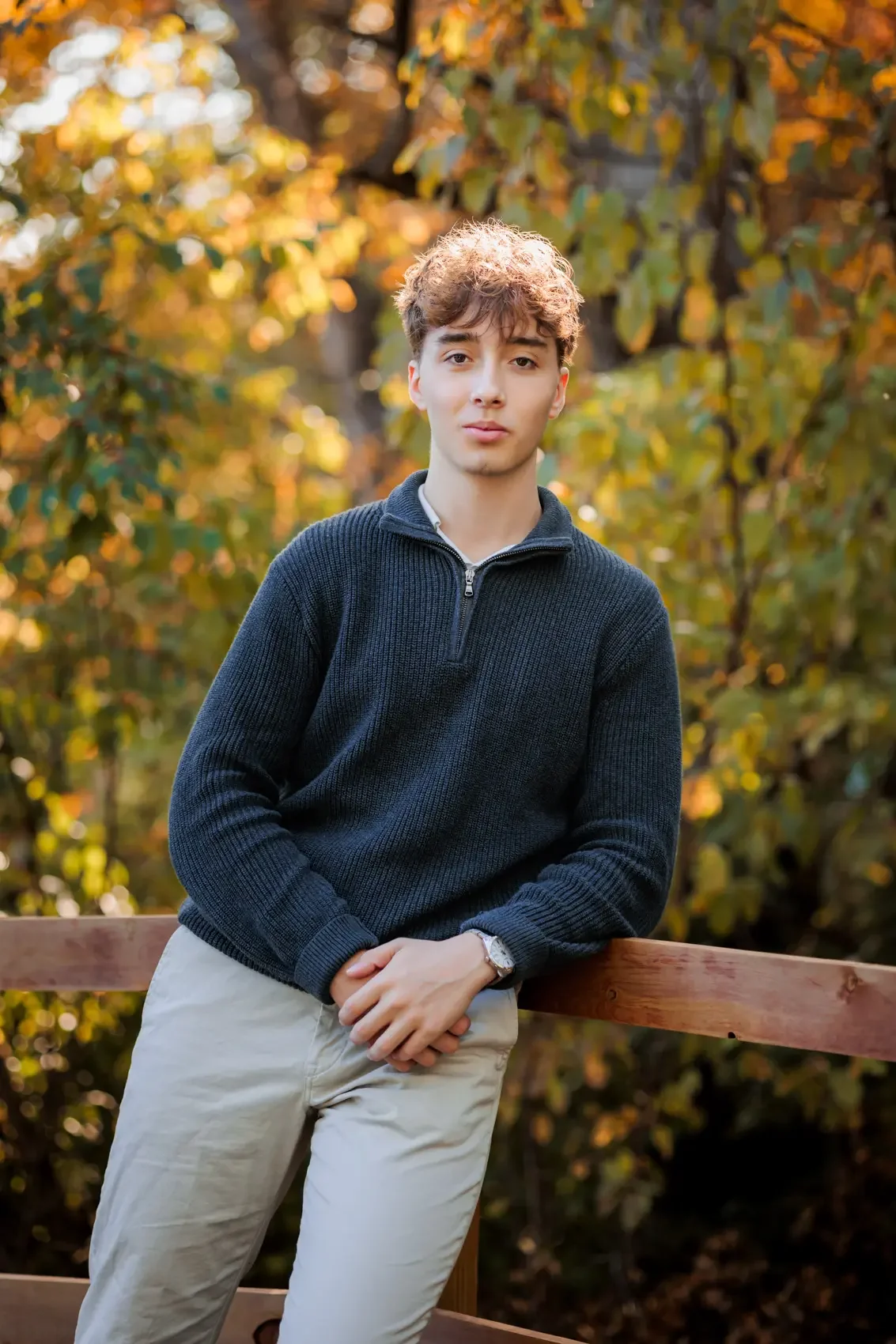 A young man with curly hair and fair skin standing outdoors in front of autumn foliage, wearing a black sweater, khaki pants, and a wristwatch, leaning against a wooden railing.