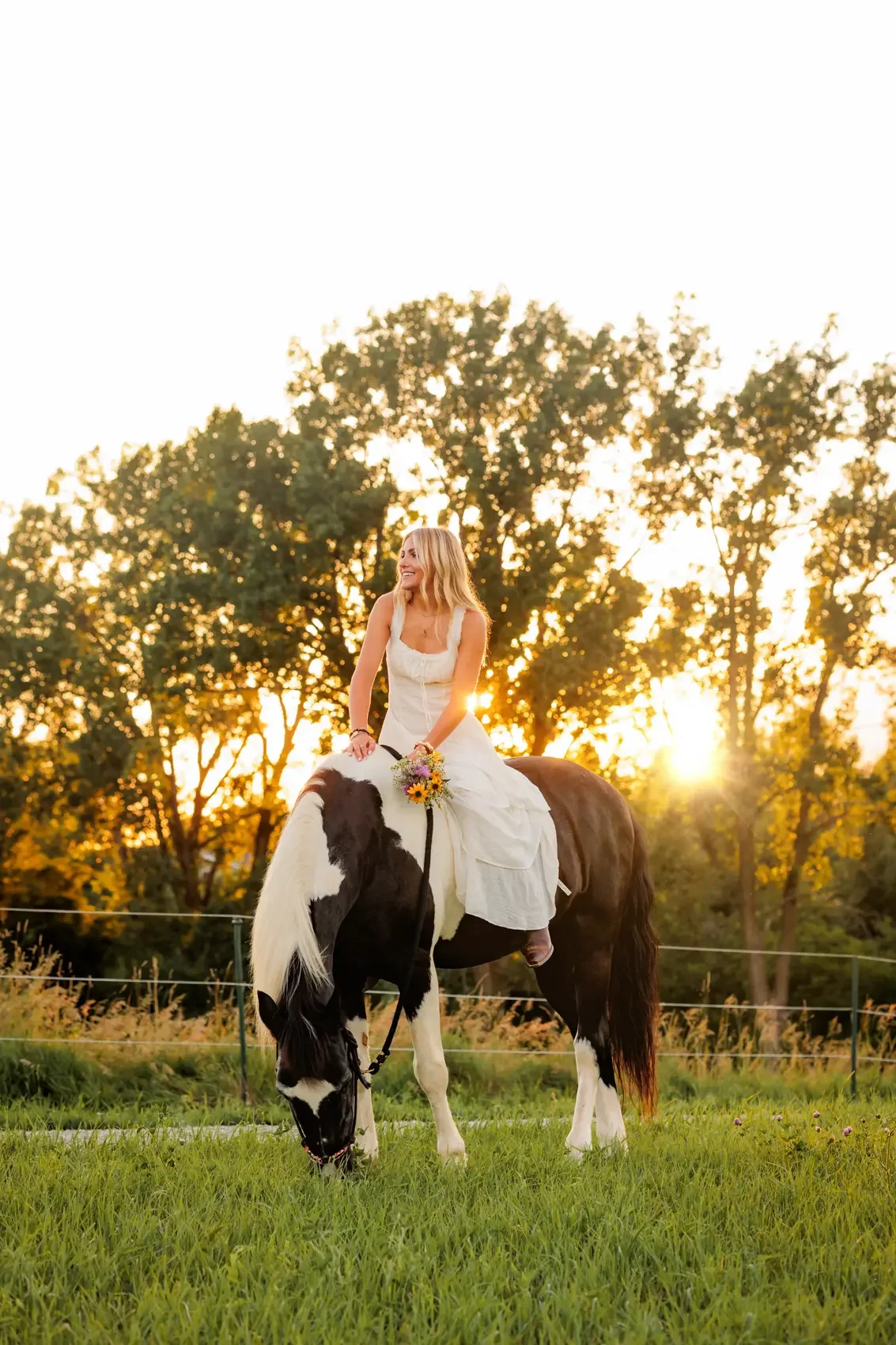 Senior girl in a white dress sitting on a black and white horse in a grassy field at sunset, holding a small bouquet of flowers and smiling.