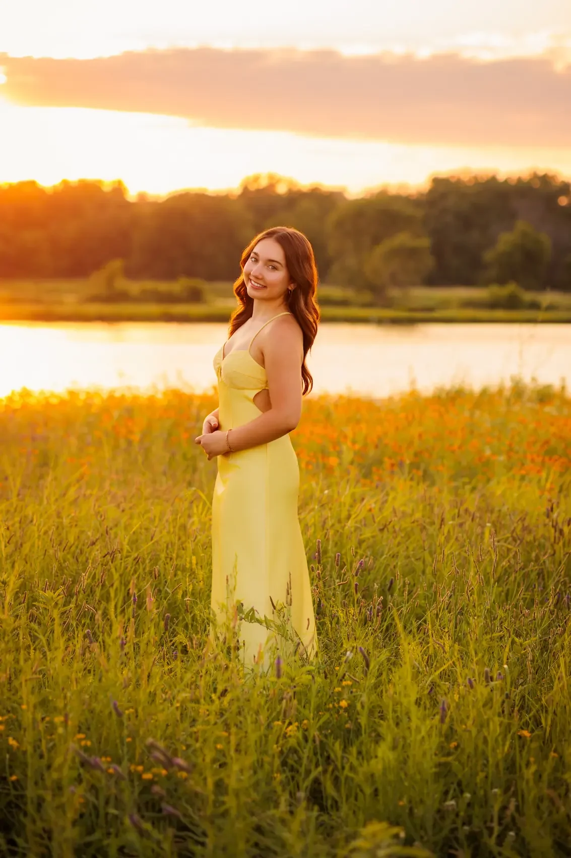 Senior girl in a yellow dress standing in a field of grass and wildflowers in Des Moines at sunset, near a body of water, smiling at the camera.