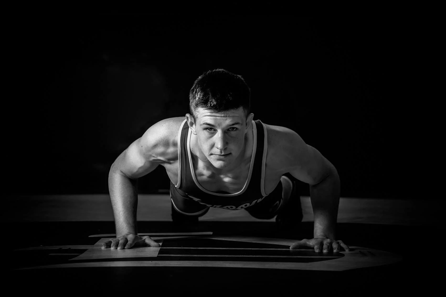 Black and white photo of a high school senior boy wrestler doing a push-up on a wooden floor with a dark background.