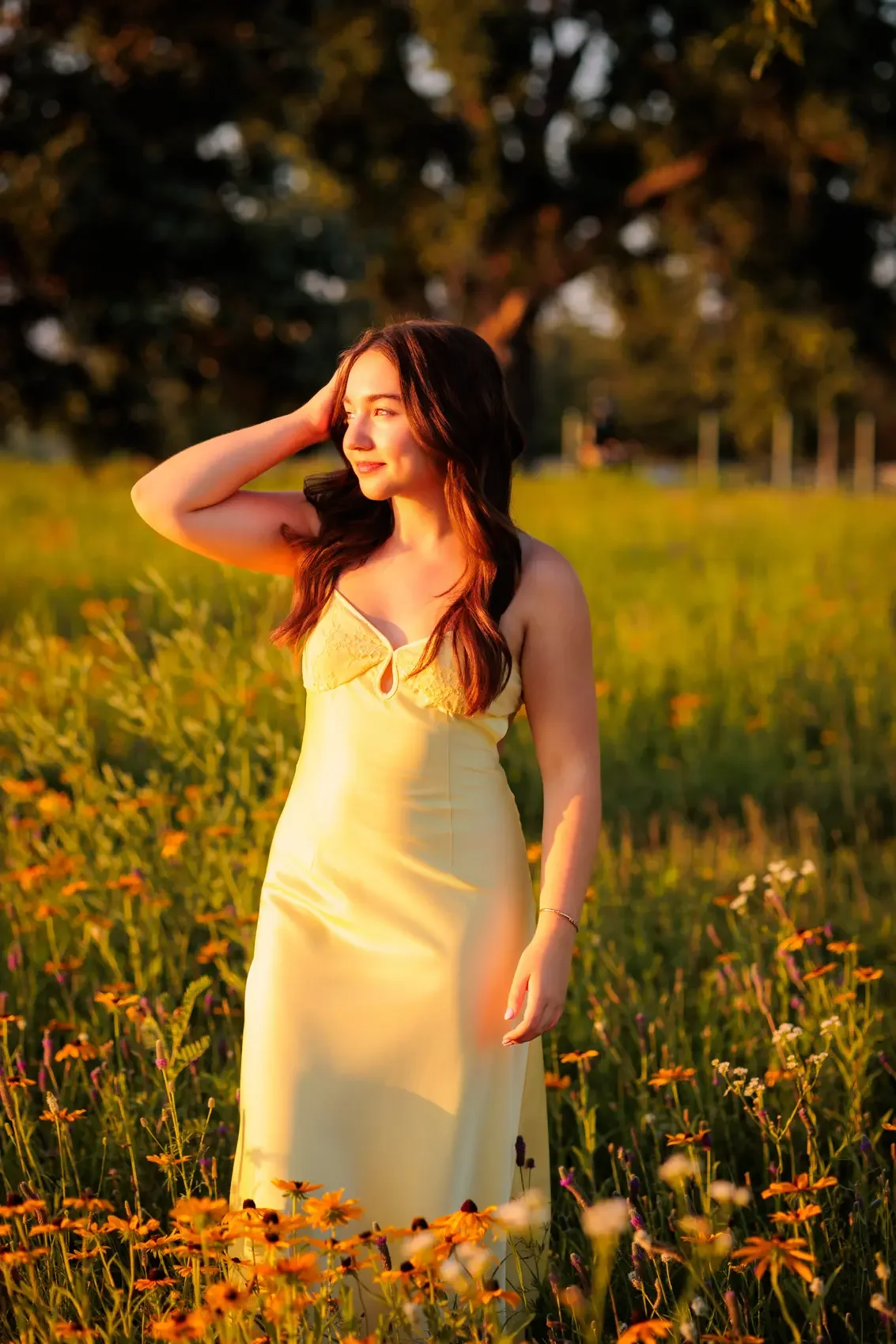 Senior girl in a yellow dress stands in a field of yellow and orange flowers during sunset, with trees in the background, looking off to the side with her hand in her hair.