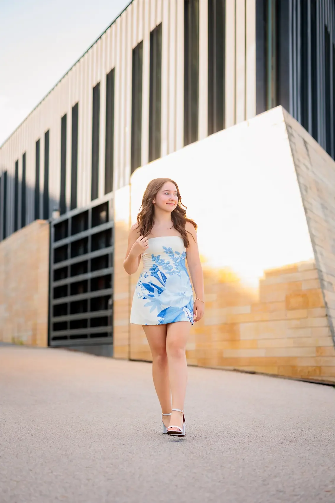 Young woman wearing a white and blue floral dress walking on a city street during sunset.