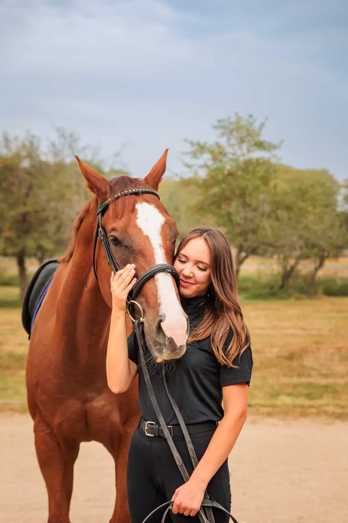Senior girl in black equestrian clothing hugging a chestnut horse with a white stripe down its face outdoors on a cloudy day with trees in the background.