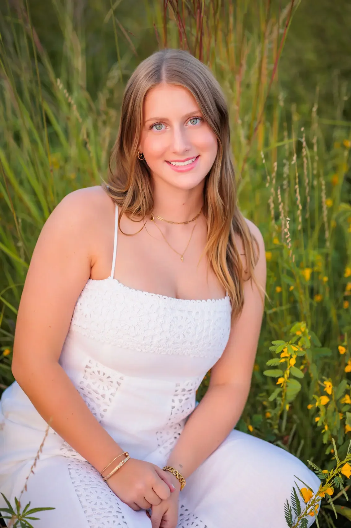 Senior girl  with long wavy brown hair and blue eyes, smiling and sitting in a field of tall green grass and yellow flowers, wearing a white sleeveless dress with lace details and gold jewelry.