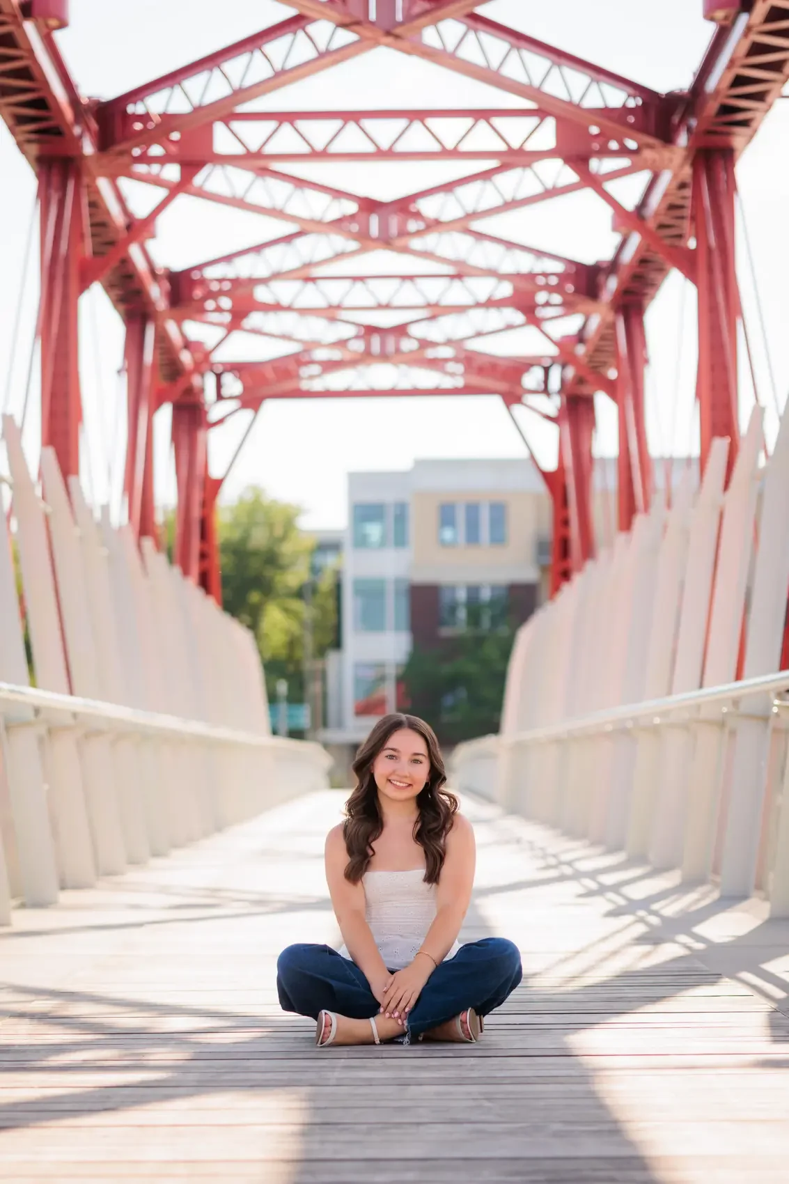 Senior girl sitting cross-legged on a wooden bridge, smiling, with a red metal bridge structure overhead and modern buildings in the background in Des Moines.