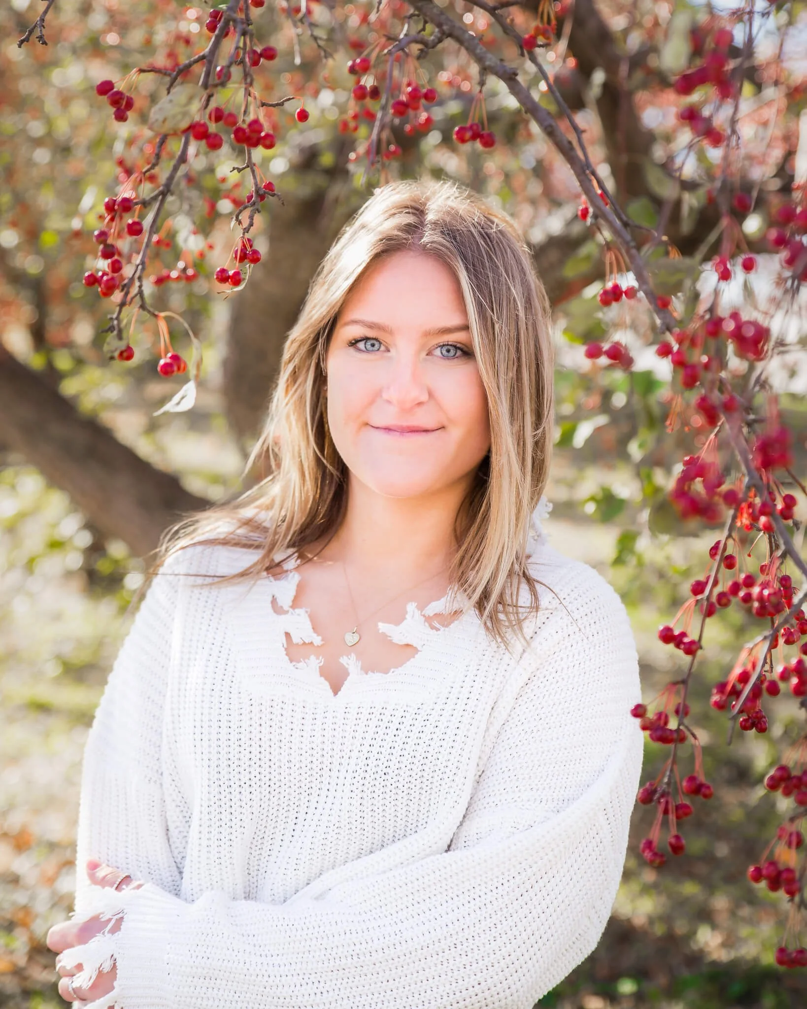 Senior girl wearing white sweater with arms crossed by colorful tree with red berries.