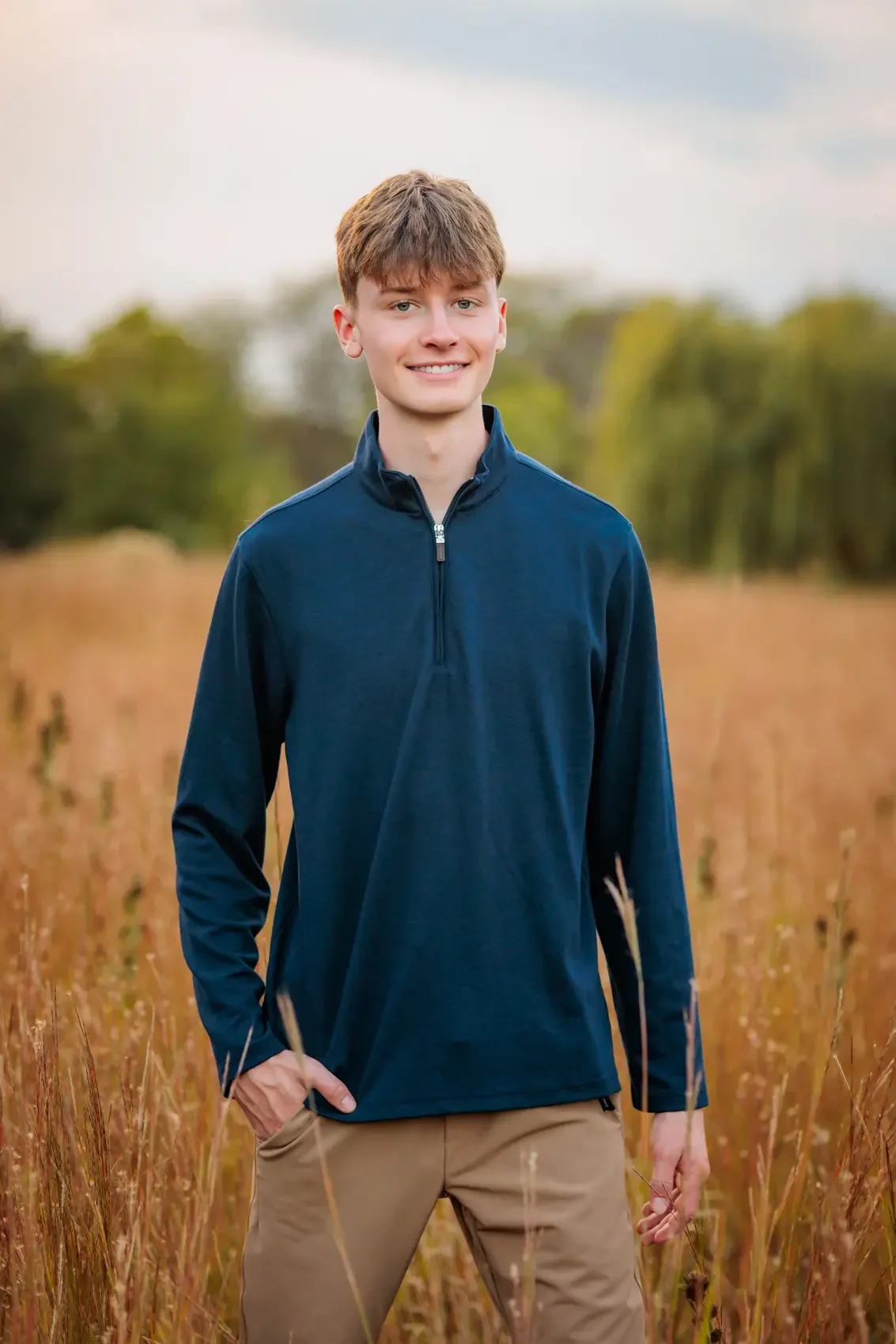 Senior boy in a navy blue quarter-zip jacket and beige pants standing in a field of tall grass with trees and a cloudy sky in the background.