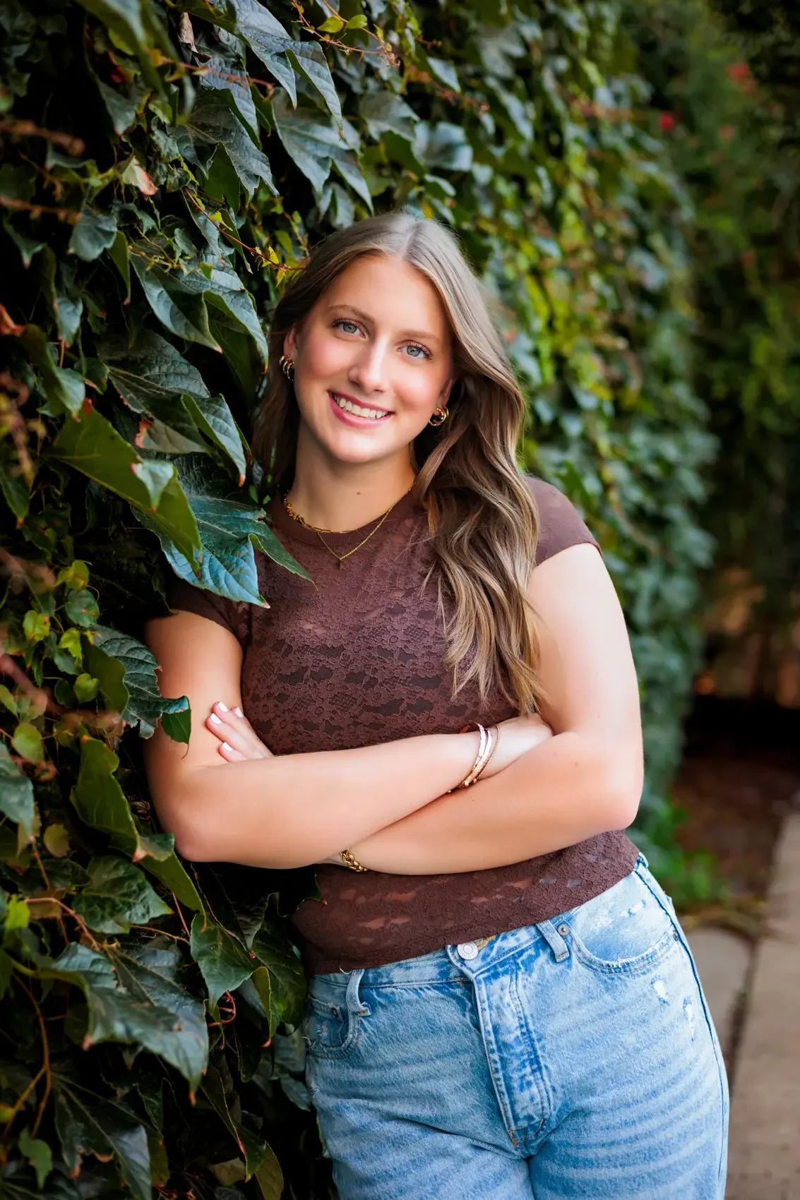 Senior girl with long wavy hair, wearing a brown lace top and light blue jeans, stands outdoors next to a green leafy hedge, smiling with arms crossed.