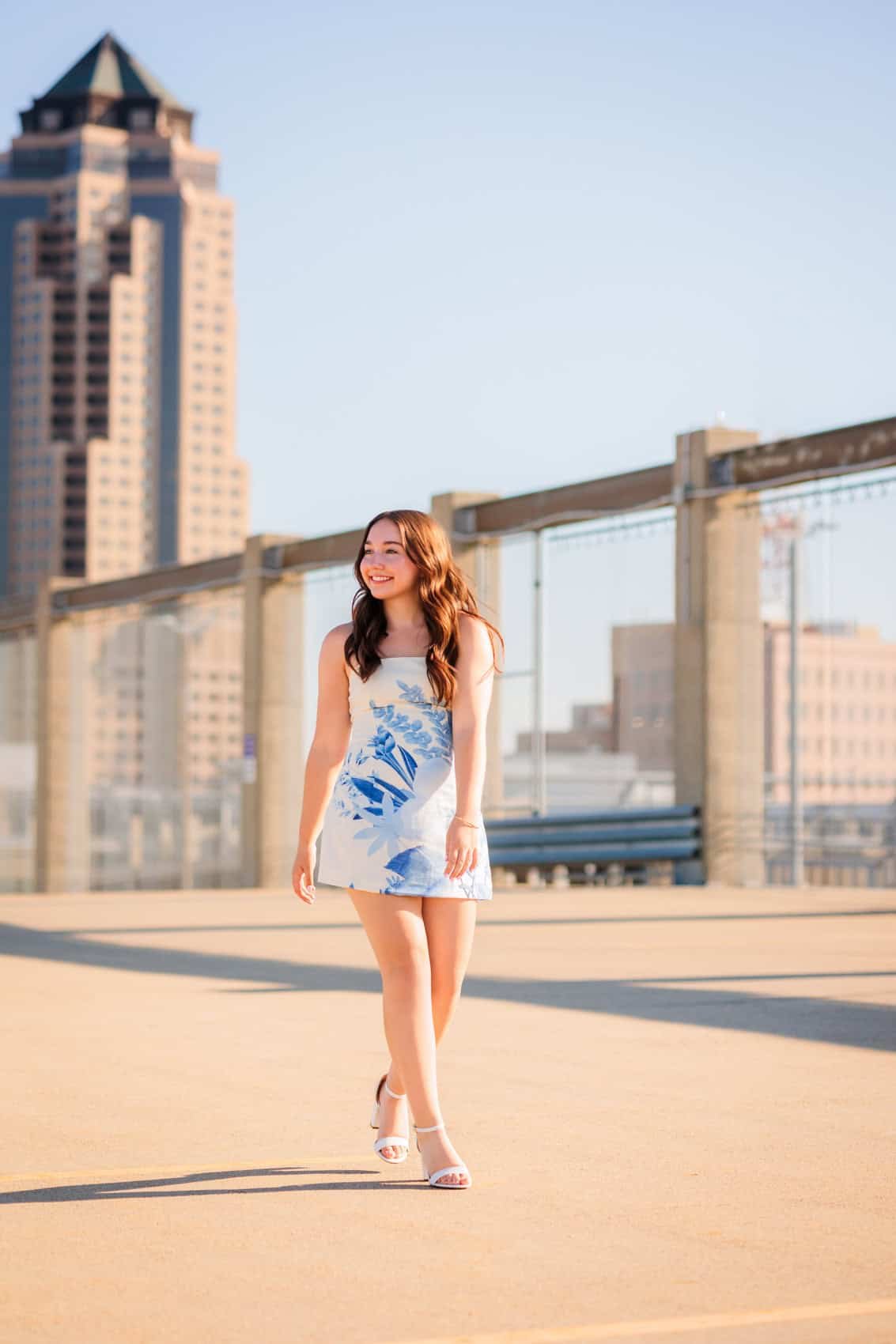  High school senior girl walking on a rooftop parking structure in Des Moines with The Principle Building in the background, wearing a white dress with blue floral patterns and white high-heeled sandals, smiling and enjoying a sunny day.