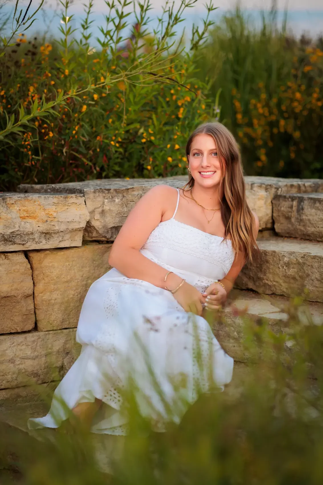 Senior girl in a white summer dress sitting on a stone wall outdoors, smiling at the camera, with green foliage and yellow flowers in the background in Des Moines.