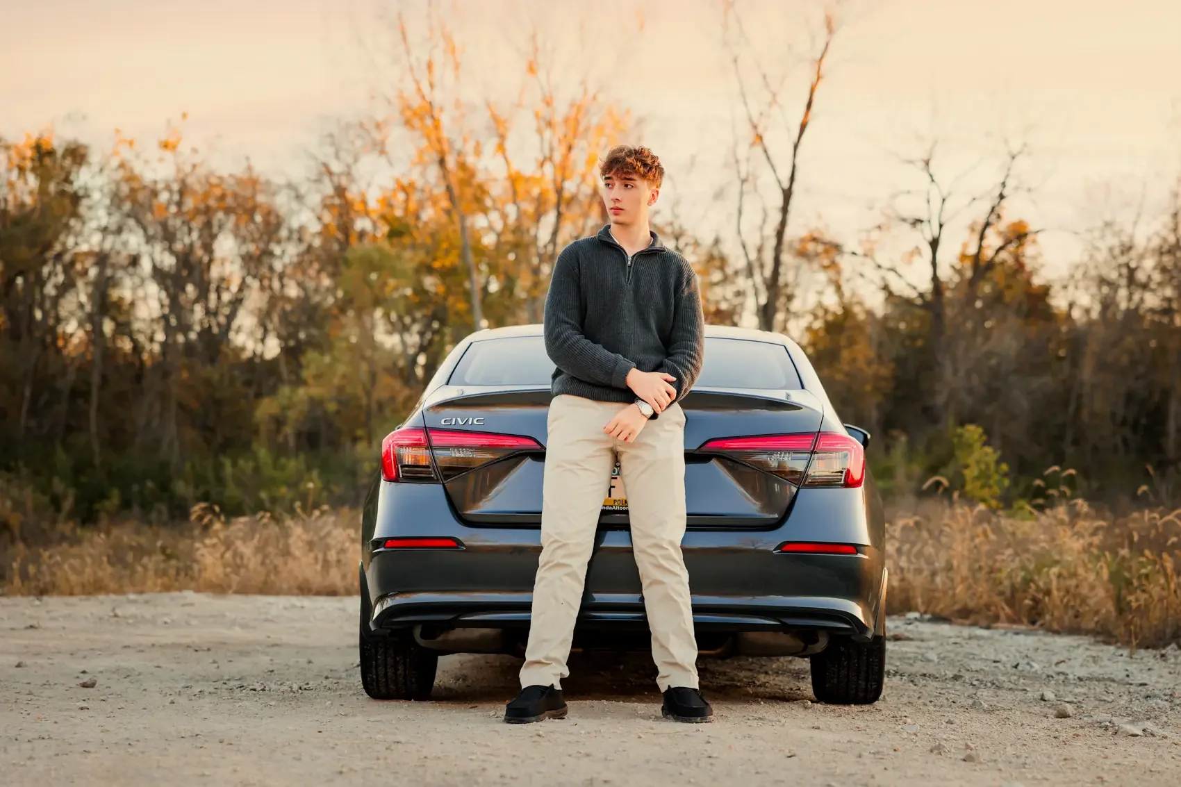 Senior boy in a dark sweater and khaki pants standing in front of a black Honda Civic car, outdoors during autumn with trees in the background.