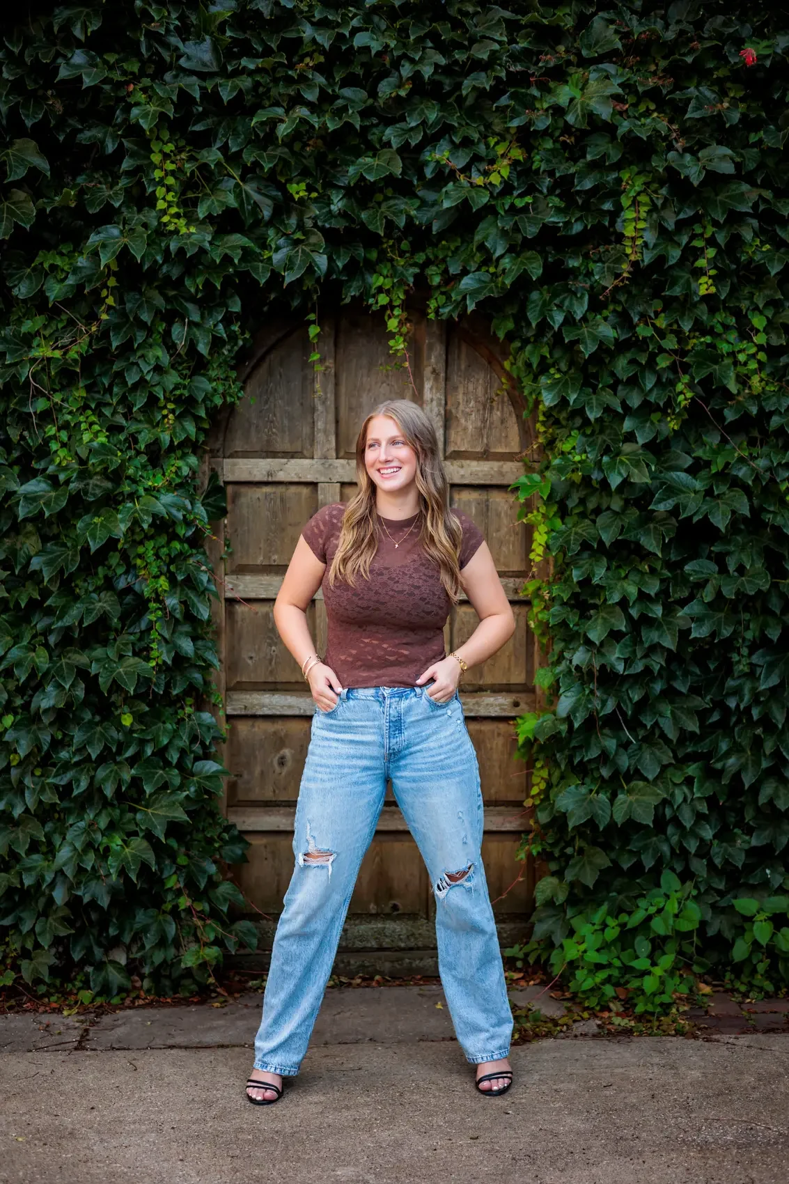 Senior girl with long wavy hair wearing a brown t-shirt, ripped jeans, and black heels standing in front of a wooden gate covered with green ivy for her senior pictures in Des Moines.