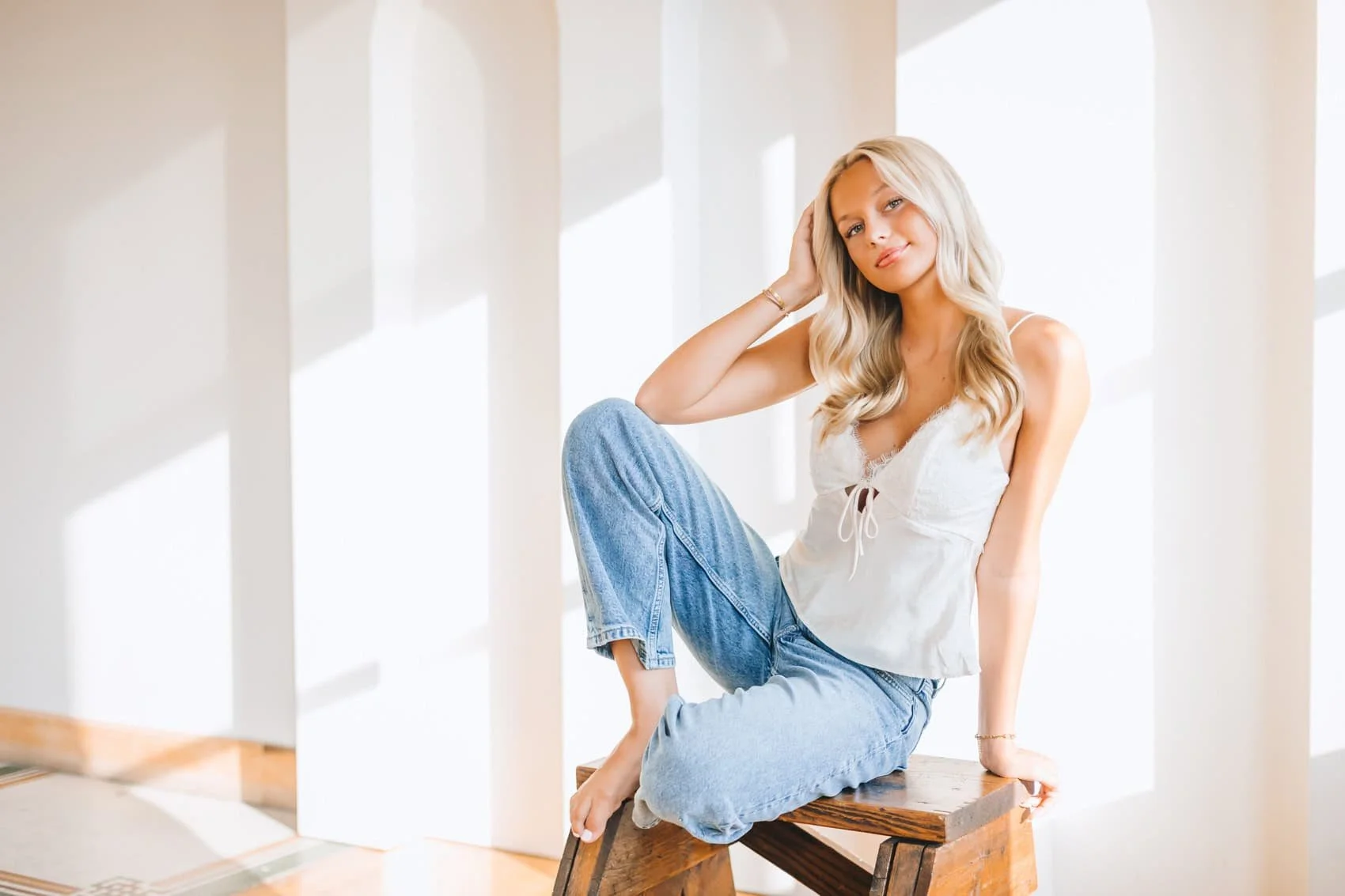 Senior girl with long blonde hair sitting on a wooden bench in a bright room with white walls and large windows, wearing a white sleeveless top and light blue jeans.
