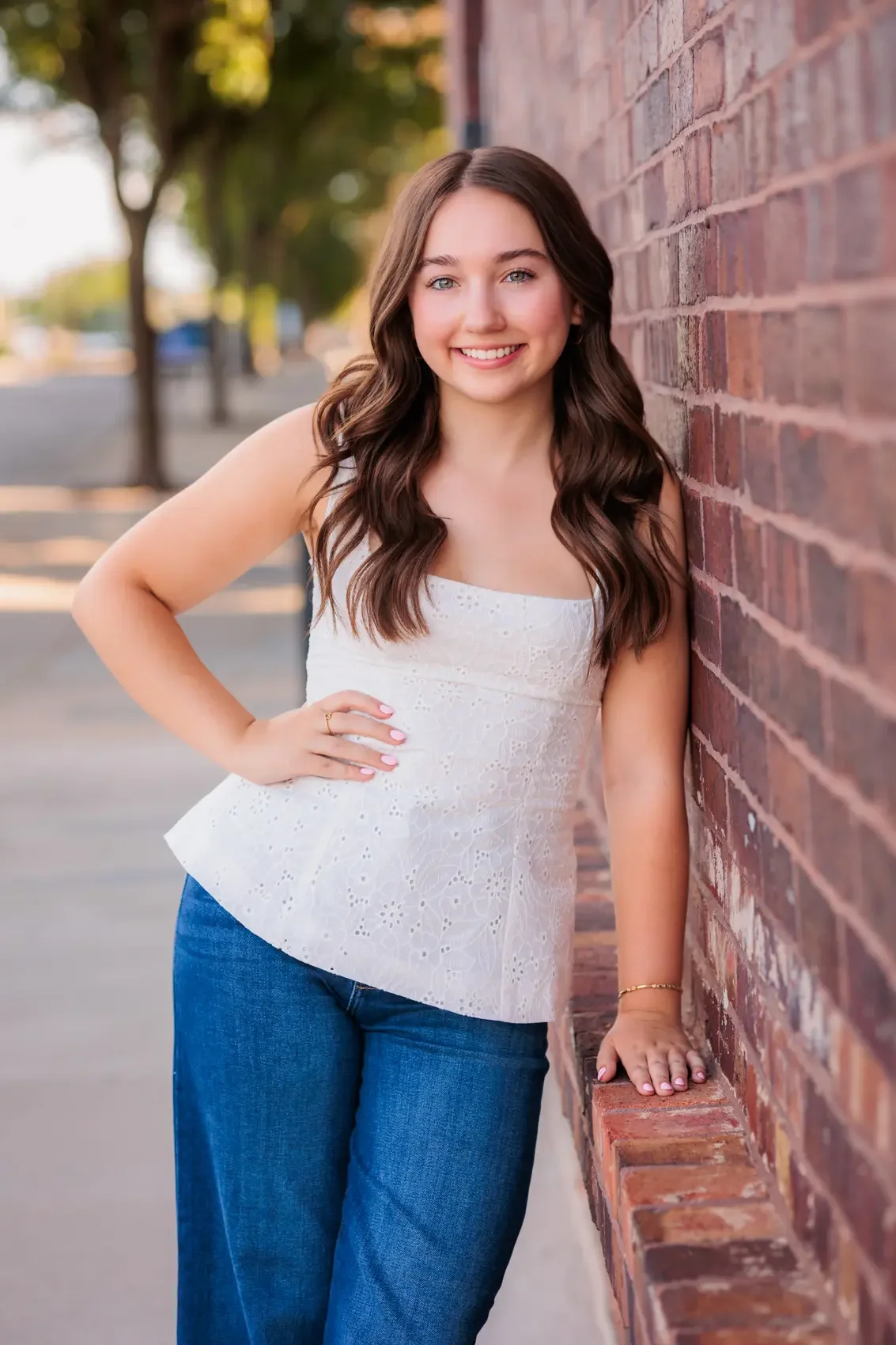 Senior girl with long, wavy brown hair and a bright smile, leaning against a brick wall outdoors during daytime in downtown Des Moines.