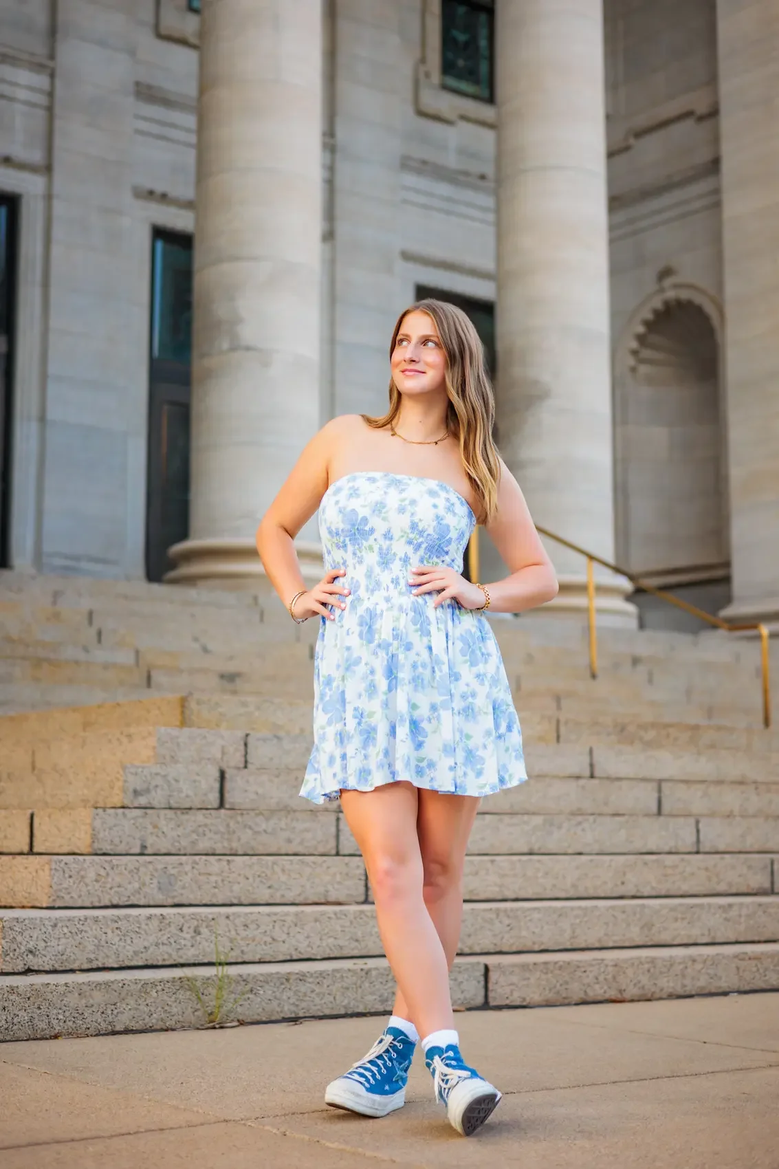 Senior girl in a strapless blue and white floral dress and blue sneakers standing on stairs outside a large stone building with columns.