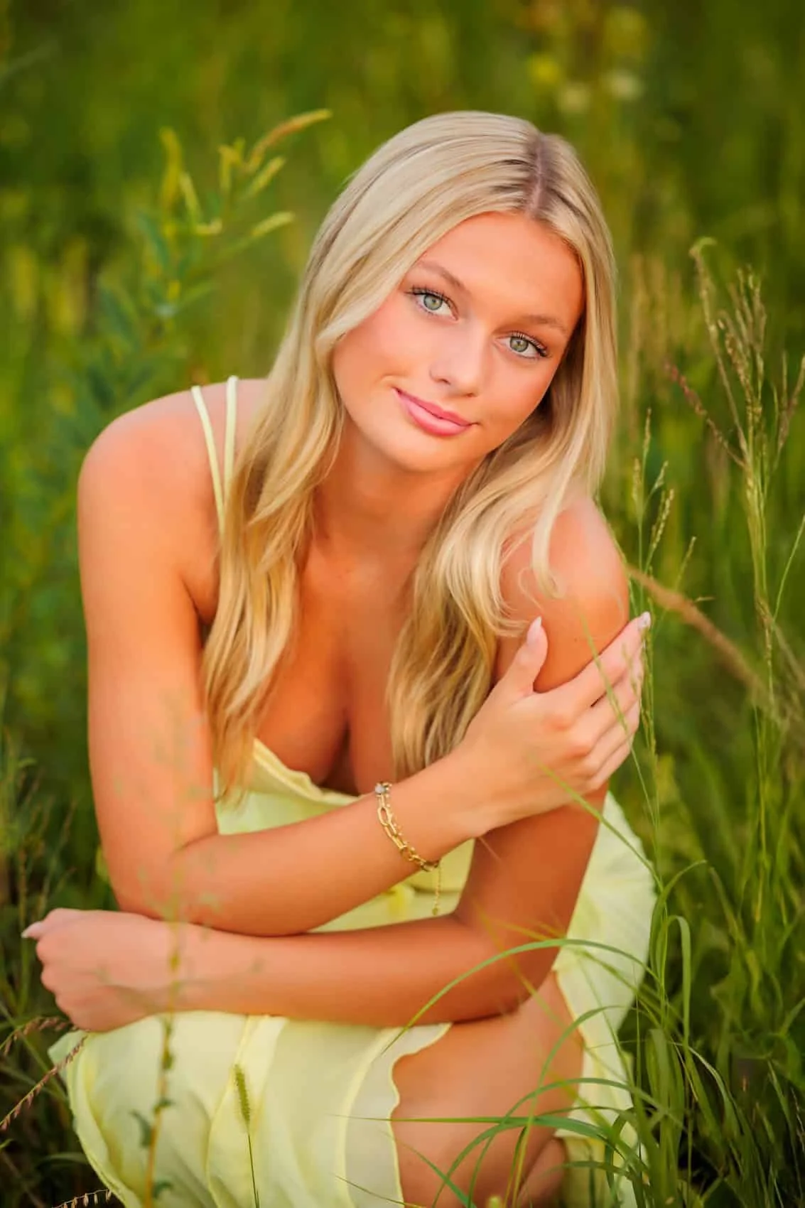 Senior girl with long blonde hair and blue eyes, wearing a yellow dress, sitting in a grassy field with green foliage, smiling gently at the camera.