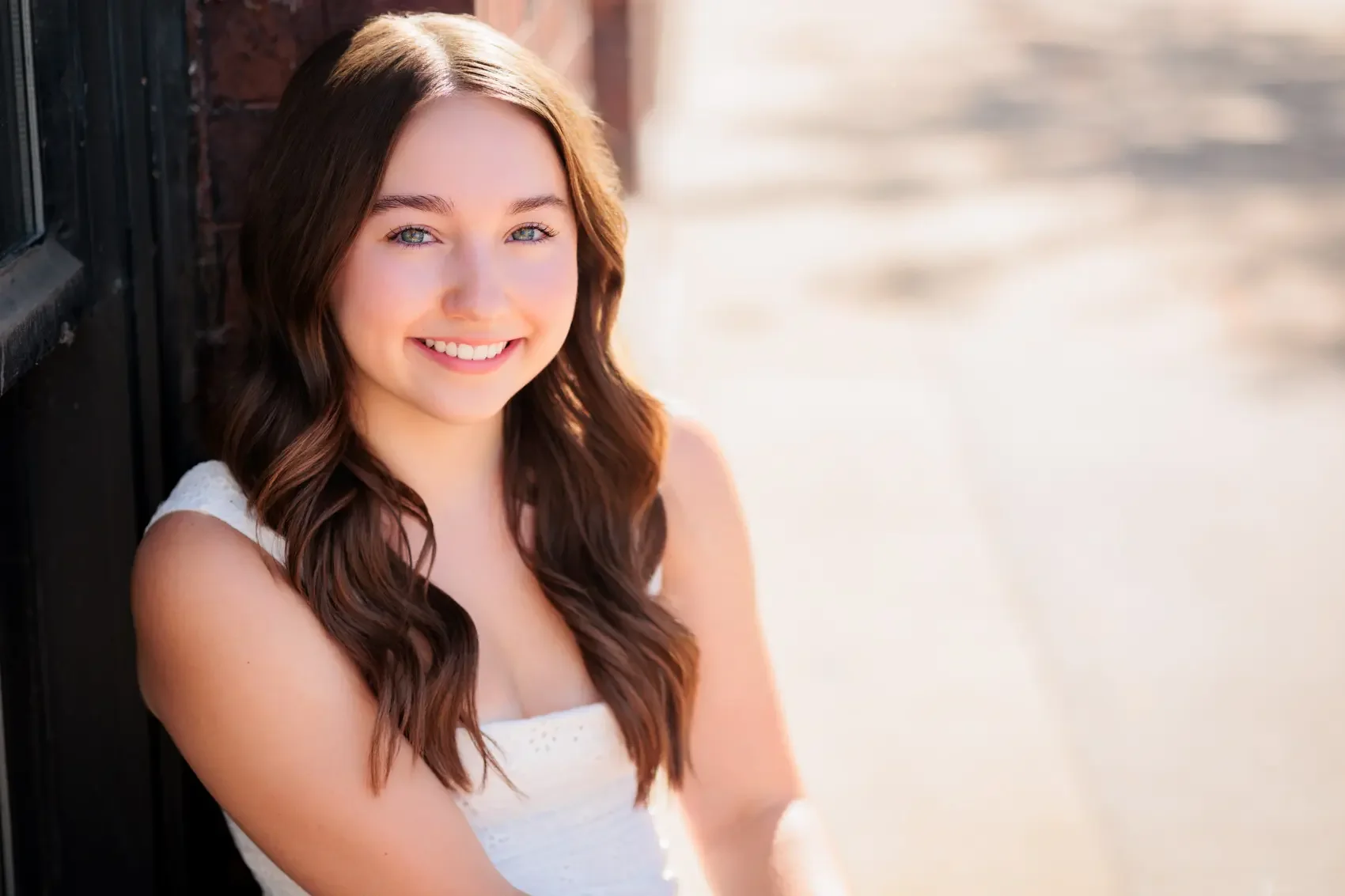 Senior girl with long wavy brown hair and blue eyes, wearing a white sleeveless top, smiling outdoors in sunlight, leaning against a brick wall in the Court Avenue district of Des Moines, Iowa.