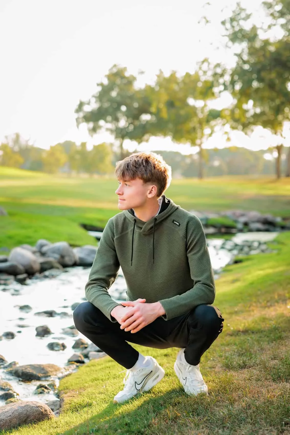  High school senior boy in a green hoodie squatting on grass near a small stream at Des Moines Golf & Country Club with trees and sunlight.